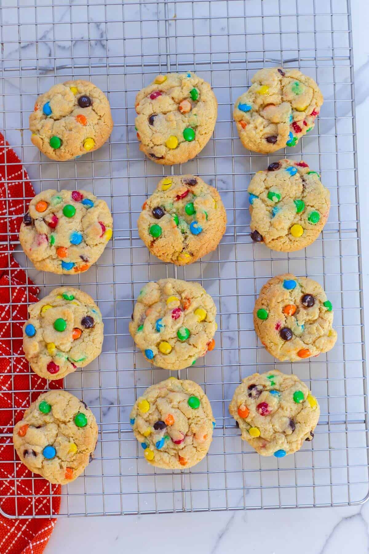 Eleven cookies with colorful candy-coated chocolates cool on a wire rack placed on a marble surface next to a red cloth.
