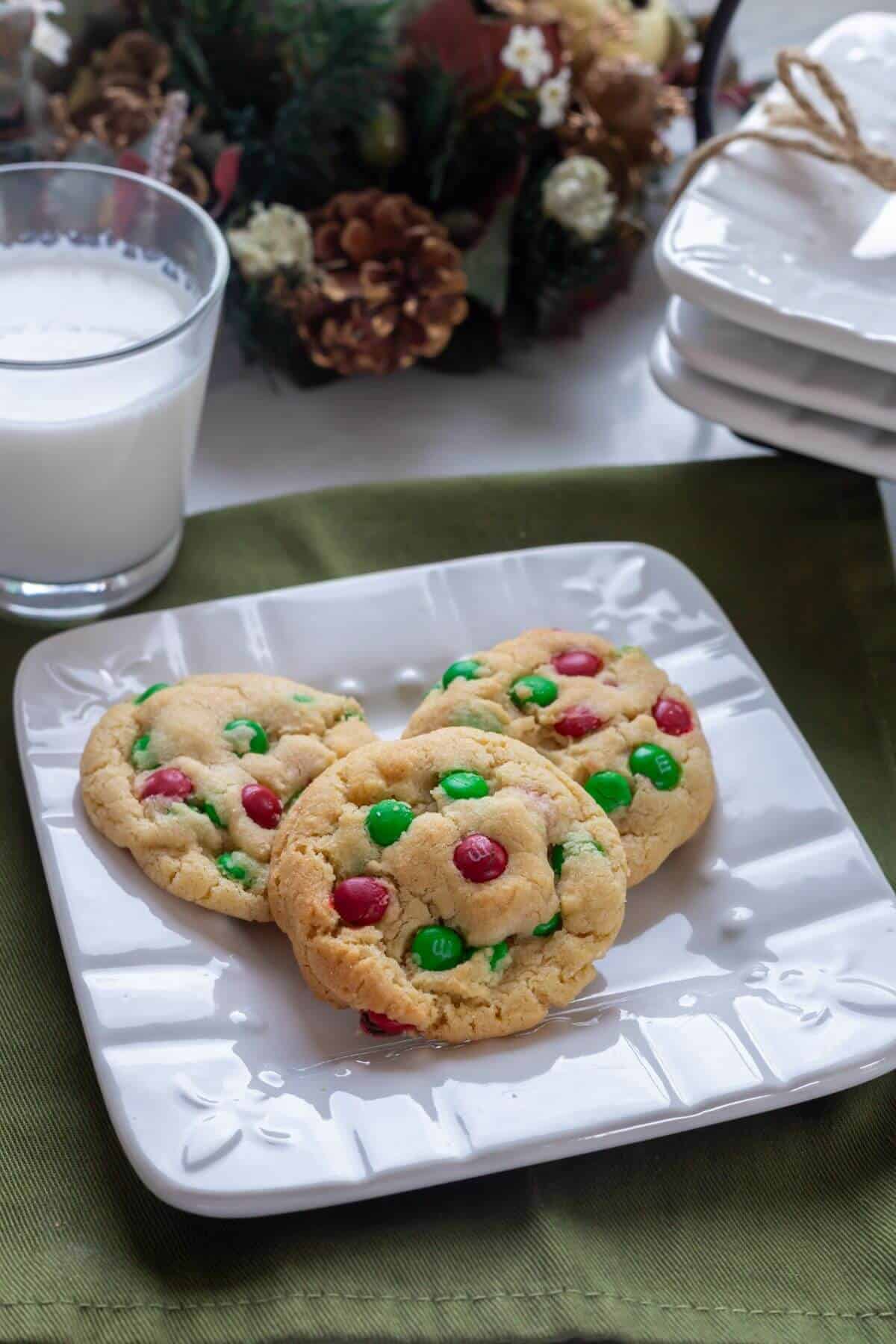 Three cookies with red and green candy pieces on a white plate, next to a glass of milk and holiday decorations in the background.