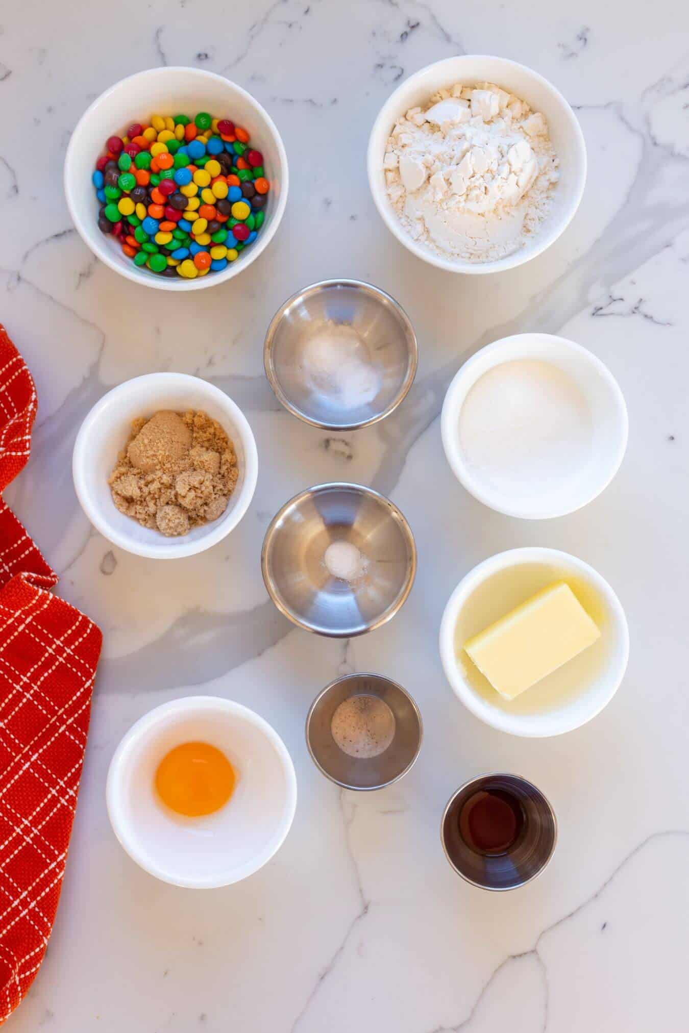 Bowls and metal cups with baking ingredients-flour, sugar, brown sugar, butter, egg yolk, mini chocolate candies, vanilla extract, and salt-arranged on a white marble surface.