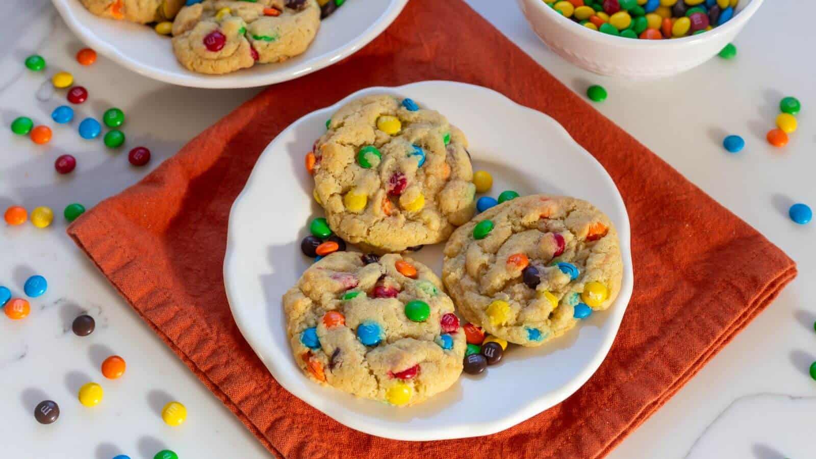Three cookies with colorful chocolate candies sit on a white plate atop a red cloth, with more cookies and candies scattered around.