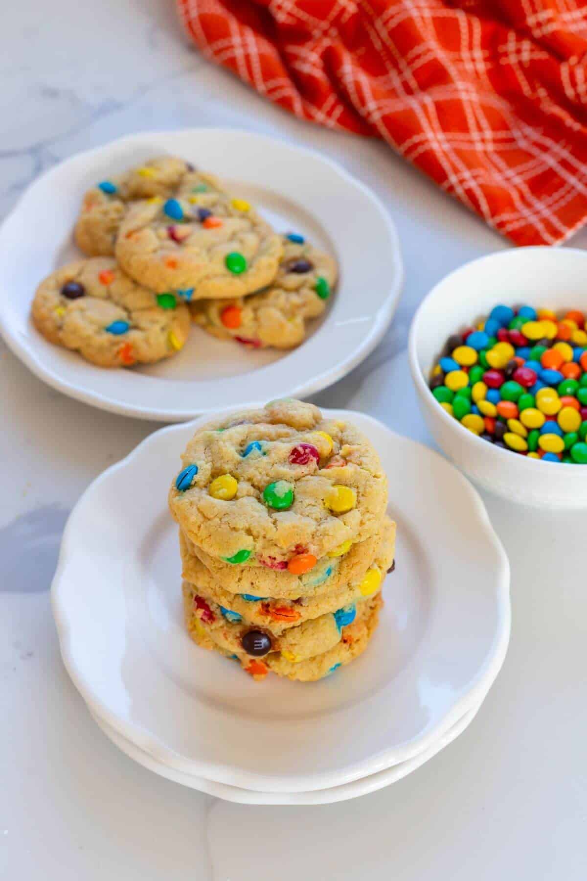 A stack of M&M cookies on a white plate, with more cookies and a bowl of M&M candies in the background on a white surface.