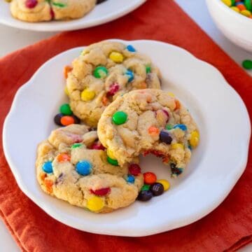 Three cookies with colorful candy-coated chocolate pieces are on a white plate, with one cookie partially eaten. The plate rests on an orange cloth.