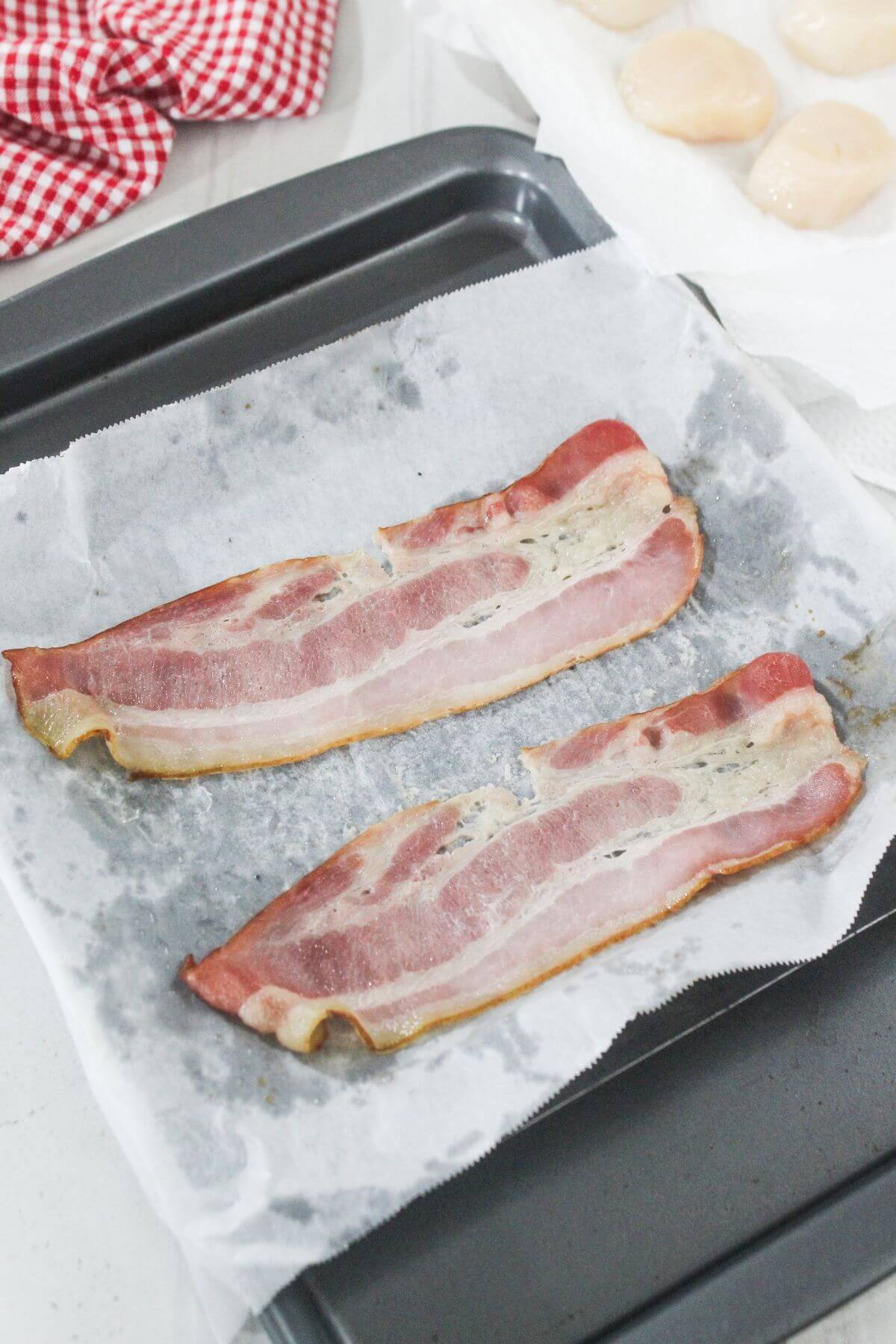 Two strips of cooked bacon on parchment paper-lined baking tray, with scallops and a red checked cloth visible in the background.