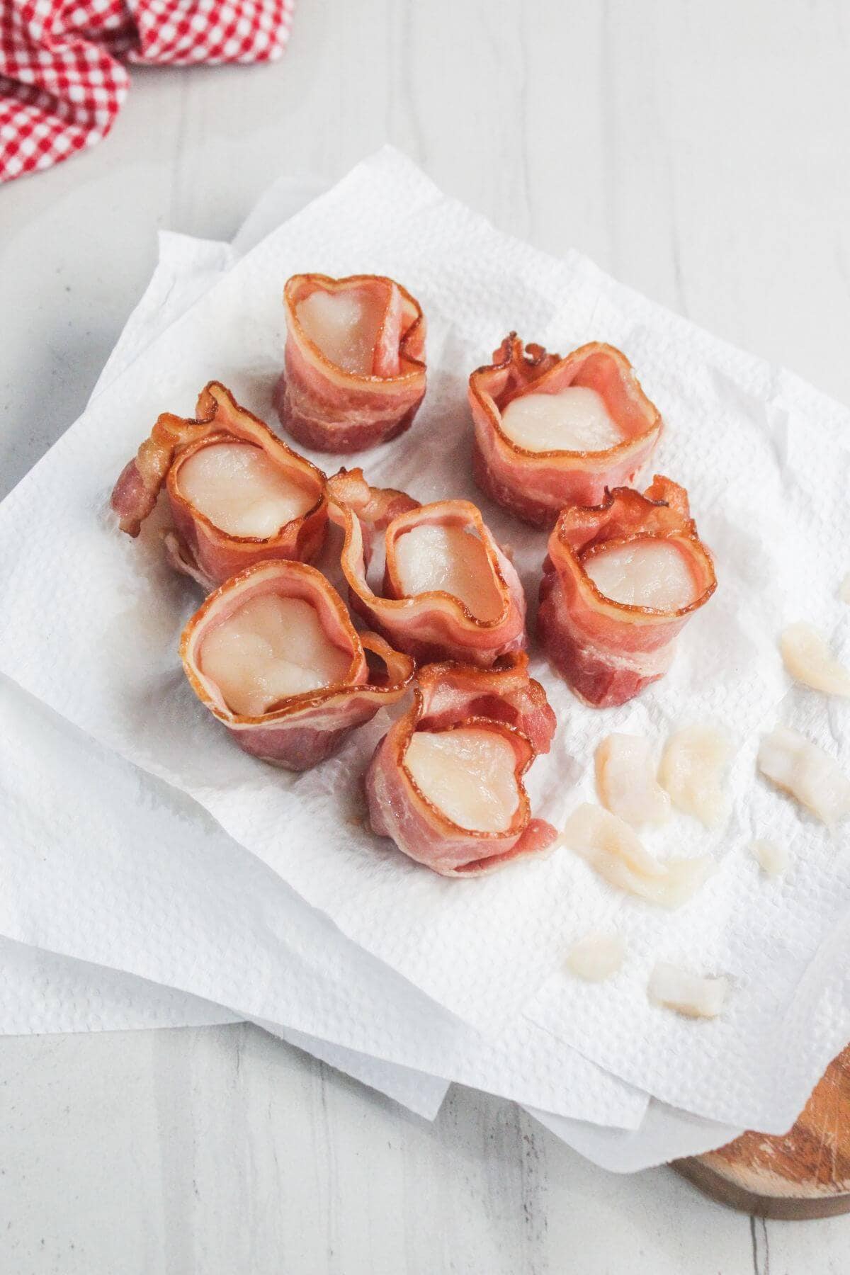 Seven pieces of bacon-wrapped scallops sit on a paper towel-lined plate, with a few small scallop pieces nearby.