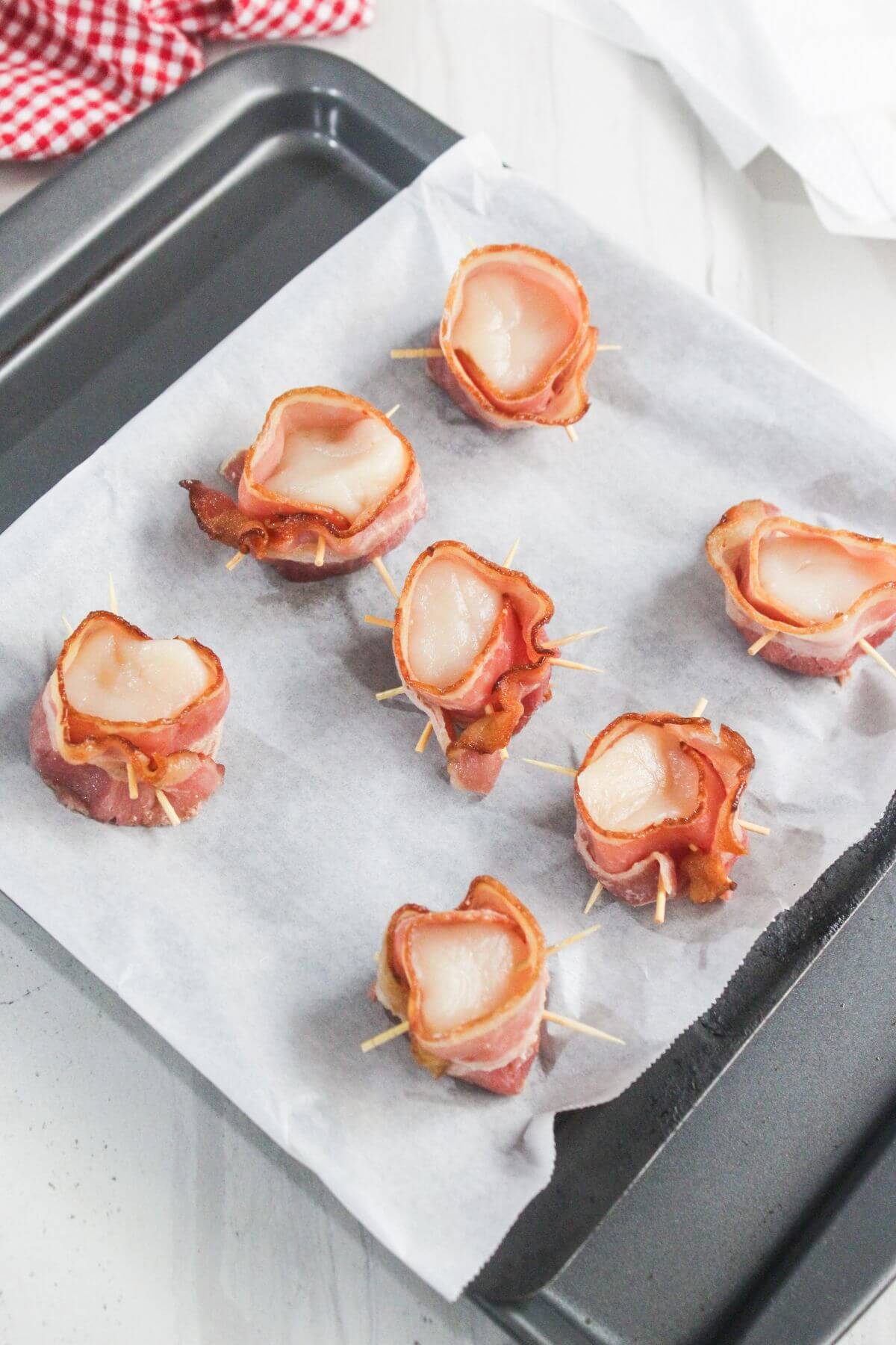 Seven bacon-wrapped scallops secured with toothpicks are arranged on a parchment-lined baking tray, ready for cooking.