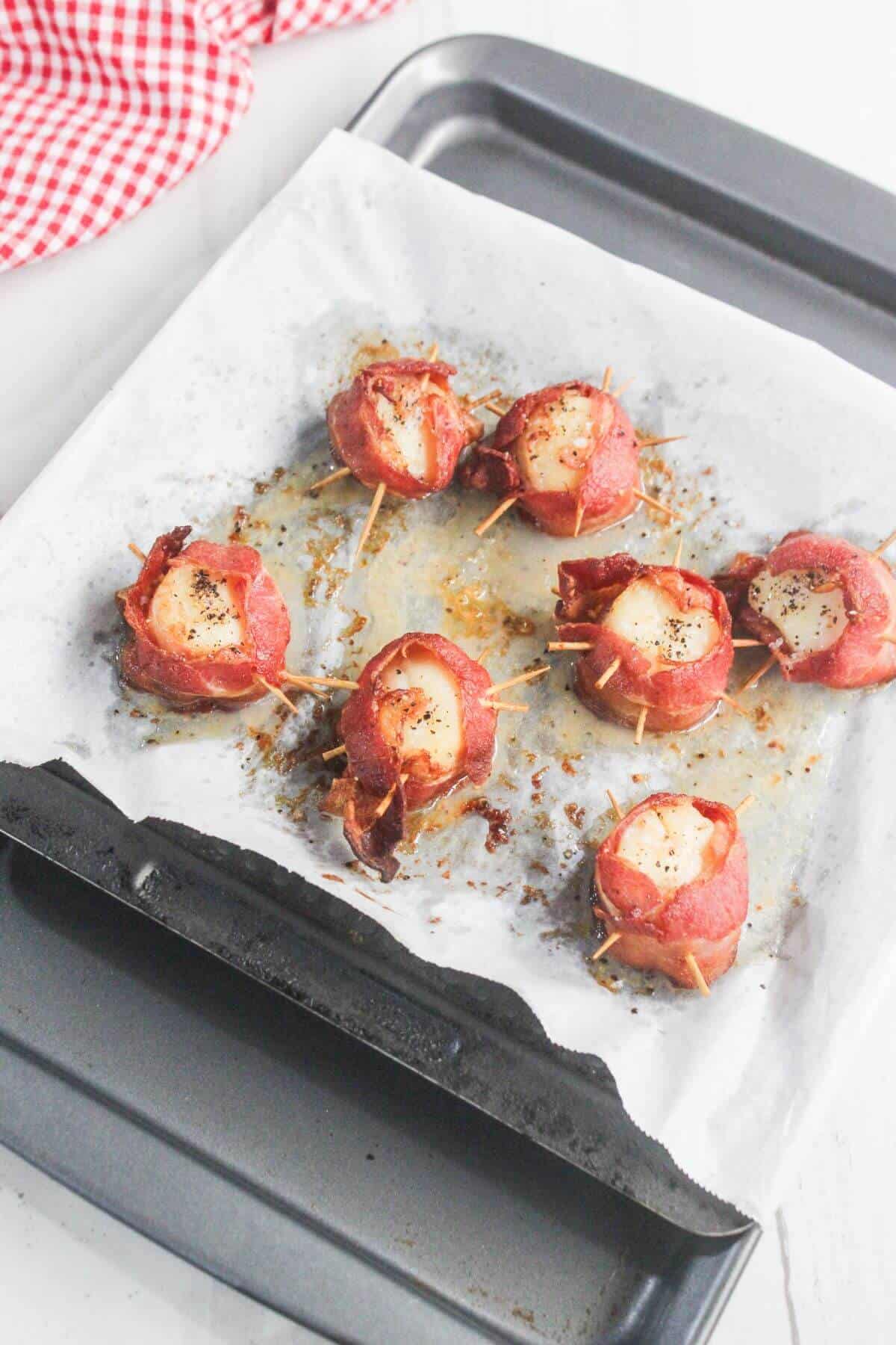 A baking tray lined with parchment paper holds seven bacon-wrapped appetizers secured with toothpicks, resting on a white surface with a red checkered cloth nearby.