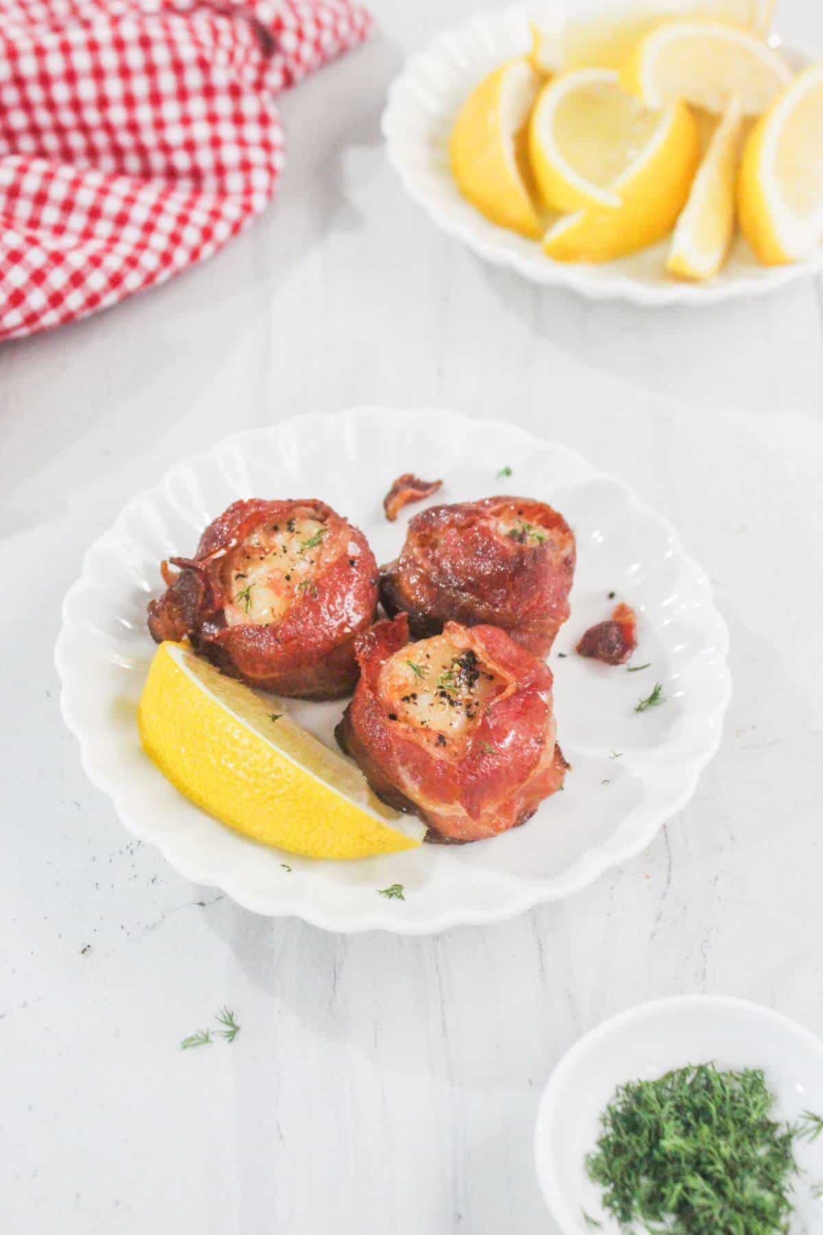 Three bacon-wrapped scallops are served on a white plate with a lemon wedge, with extra lemon slices and chopped herbs in the background.