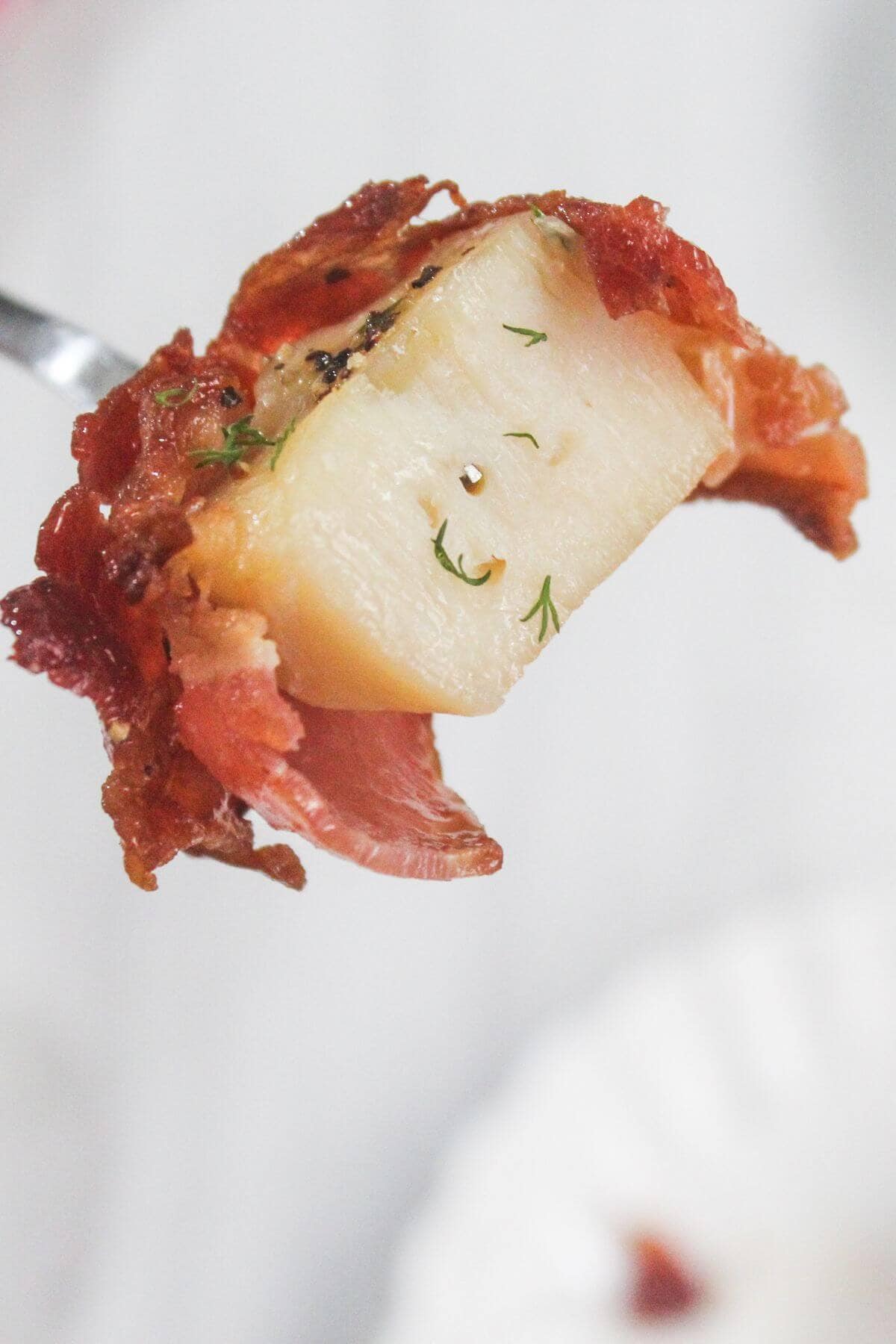 A close-up of a bacon-wrapped scallop garnished with herbs, held up on a fork against a light background.