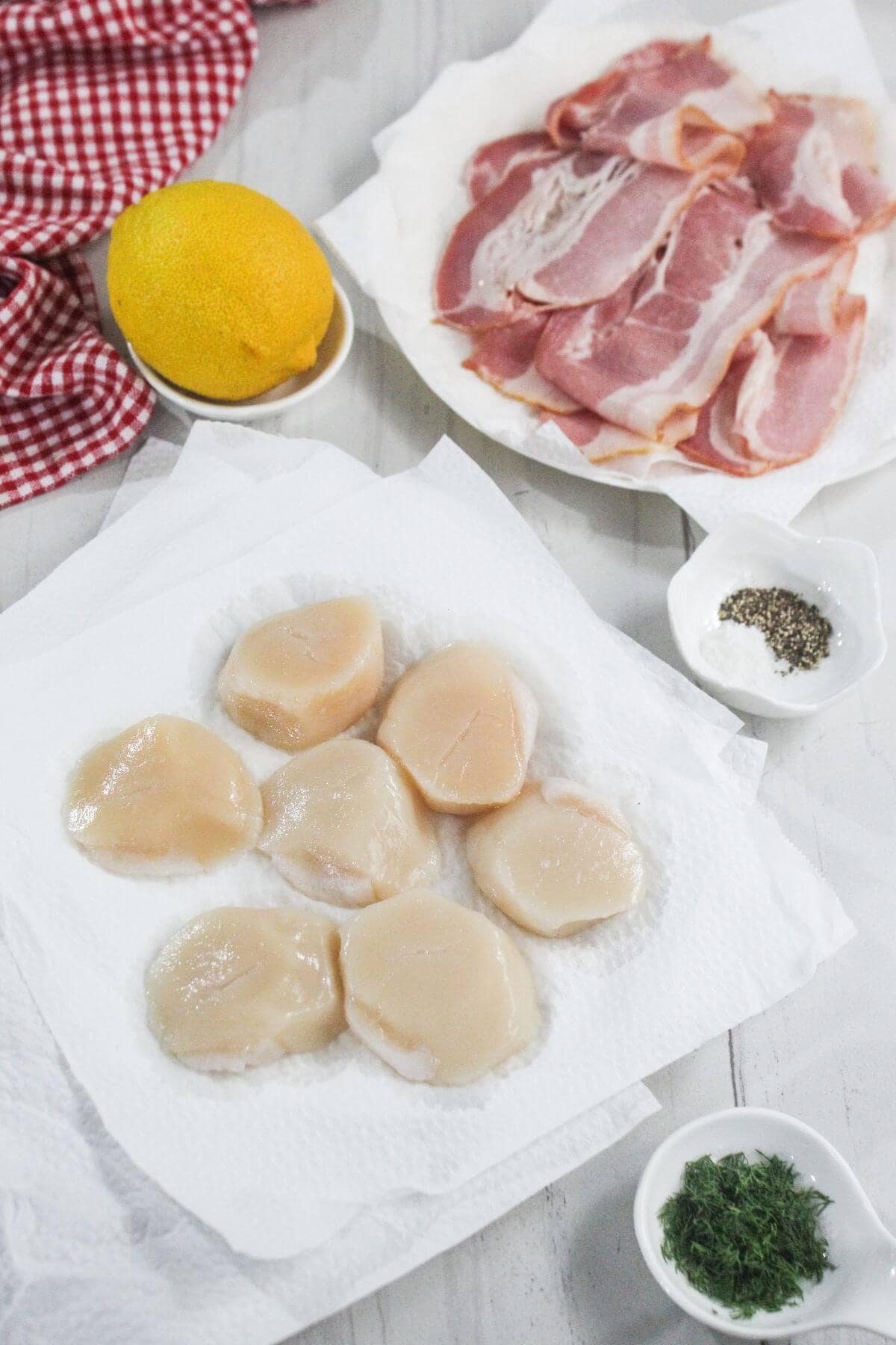 Raw scallops on a paper towel, slices of bacon on a plate, a lemon, bowls of chopped herbs, and ground black pepper on a white surface.