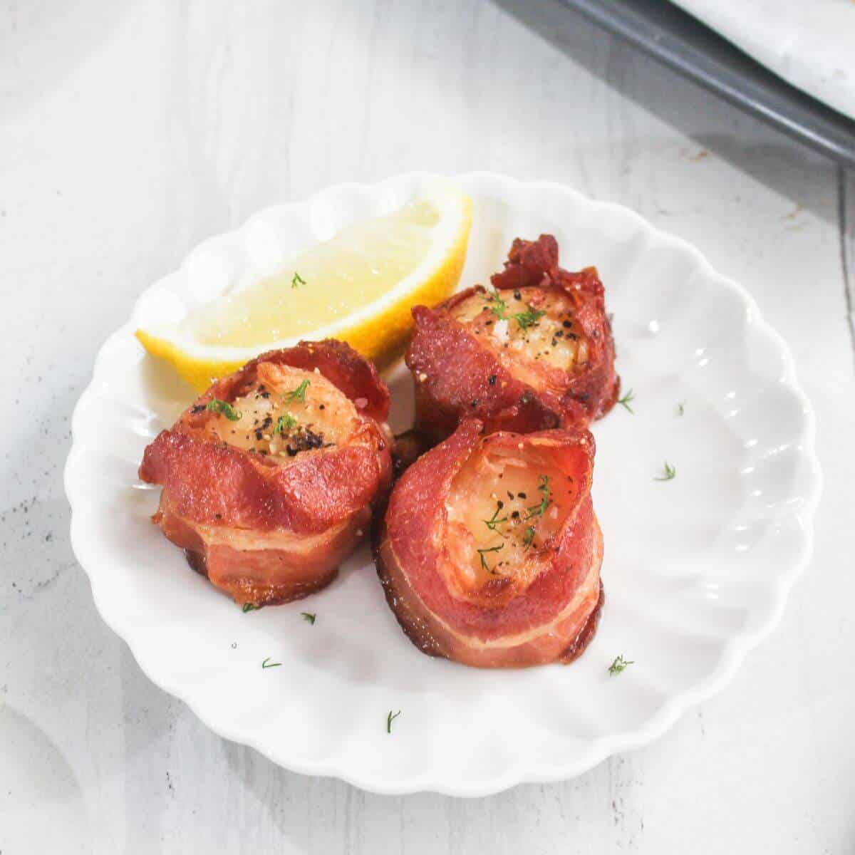 Three bacon-wrapped scallops on a white plate, garnished with herbs, served with a lemon wedge.