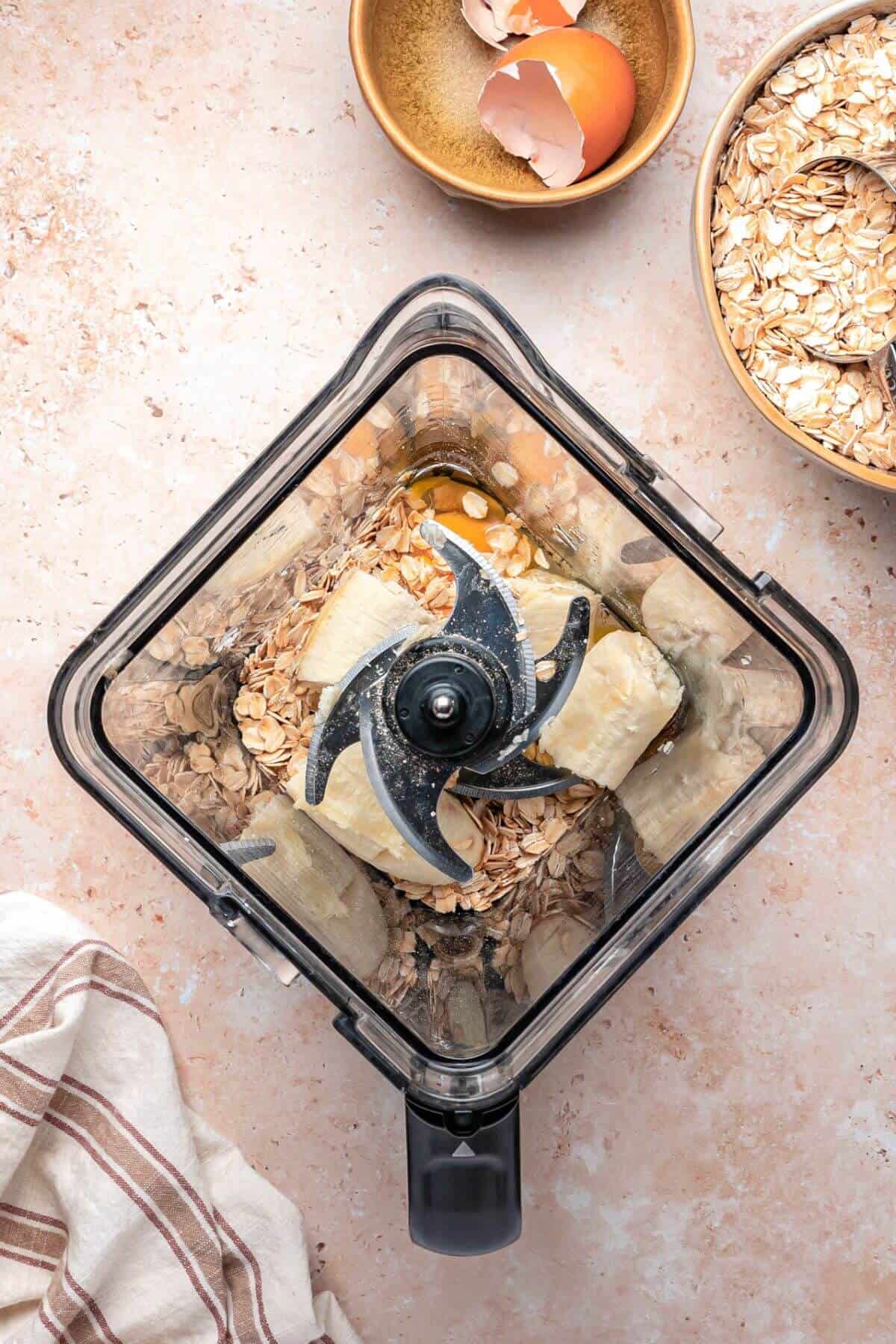 A food processor bowl containing oats, banana pieces, and seeds, viewed from above; nearby are bowls of oat flakes and eggshells.