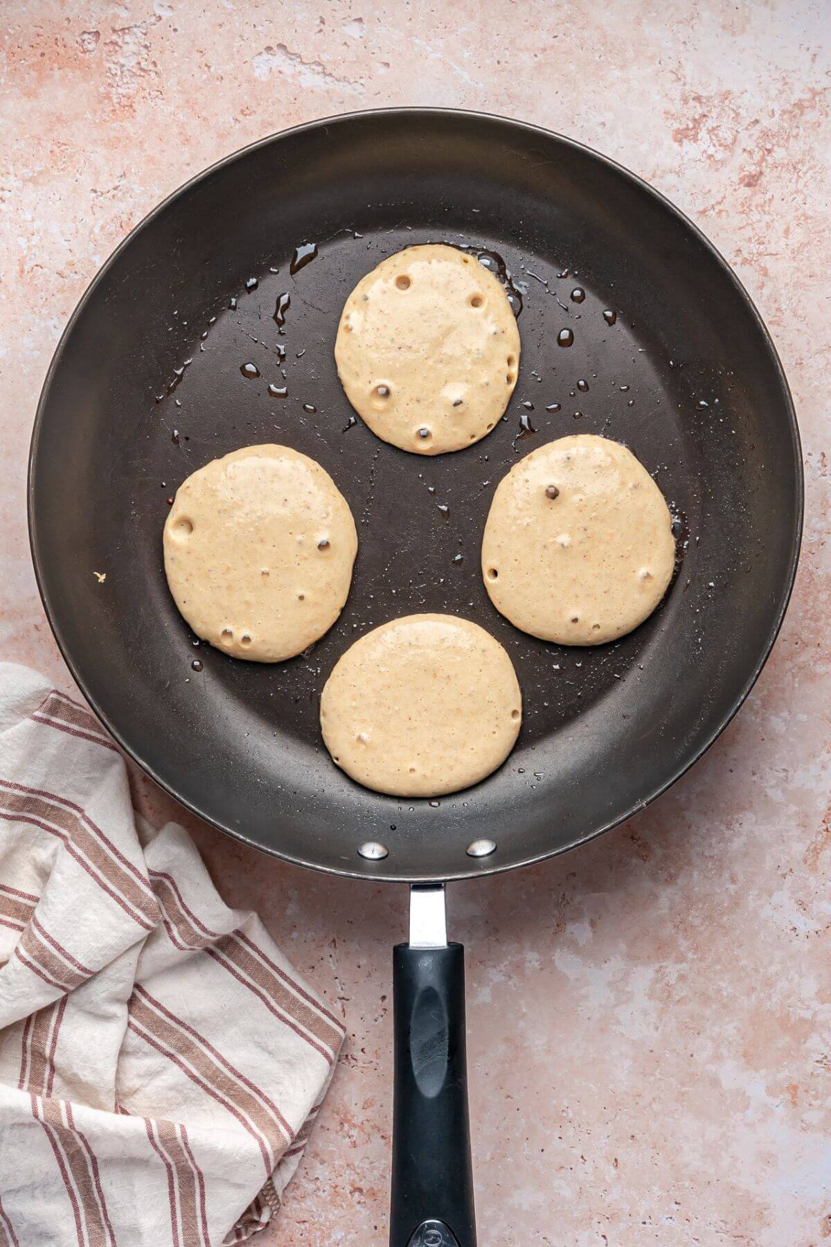 Four small pancakes are cooking in a nonstick frying pan with some oil; a striped kitchen towel is beside the pan on a light countertop.