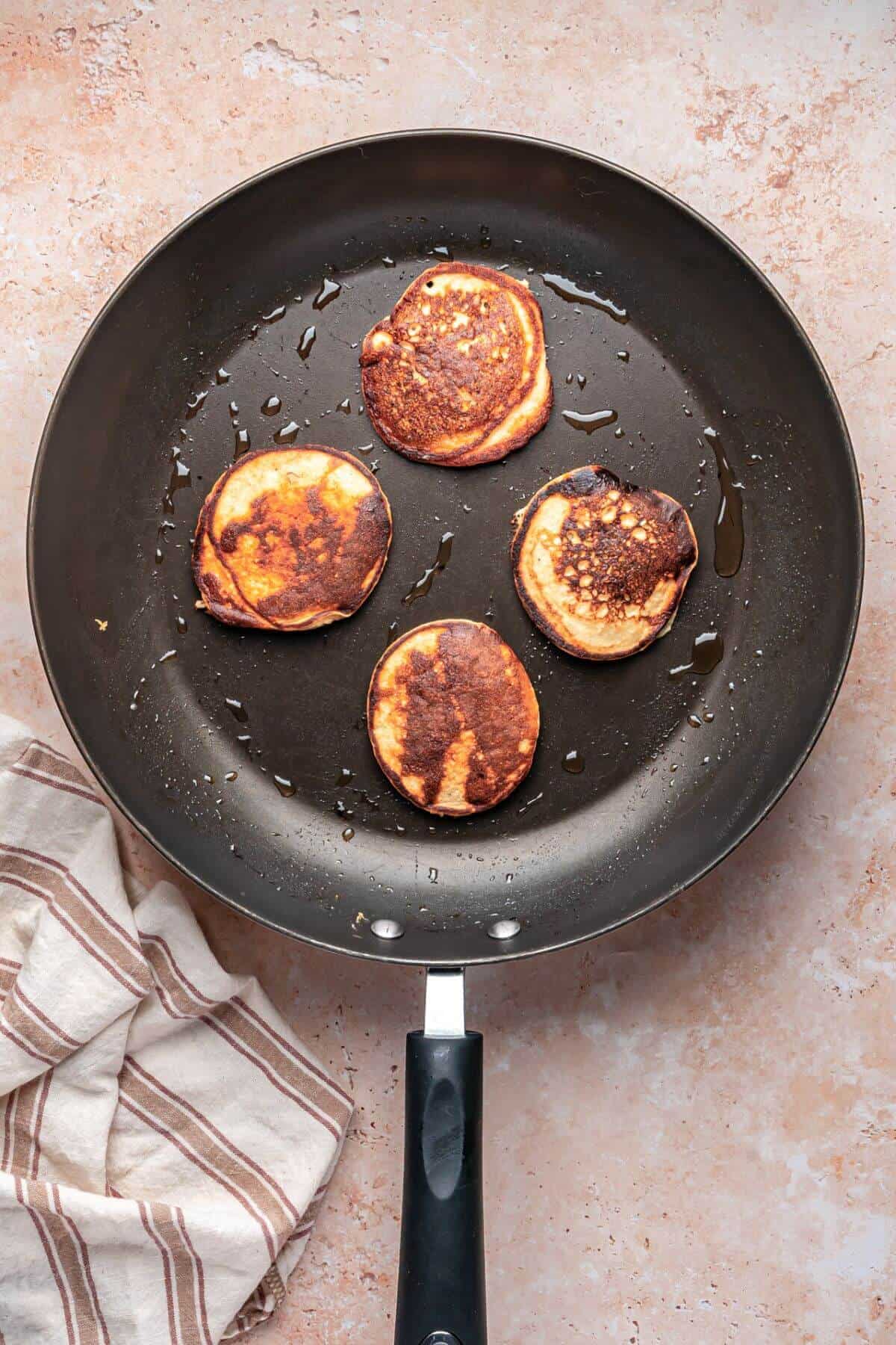 A black frying pan with four small, browned pancakes sits on a light-colored surface next to a striped kitchen towel.