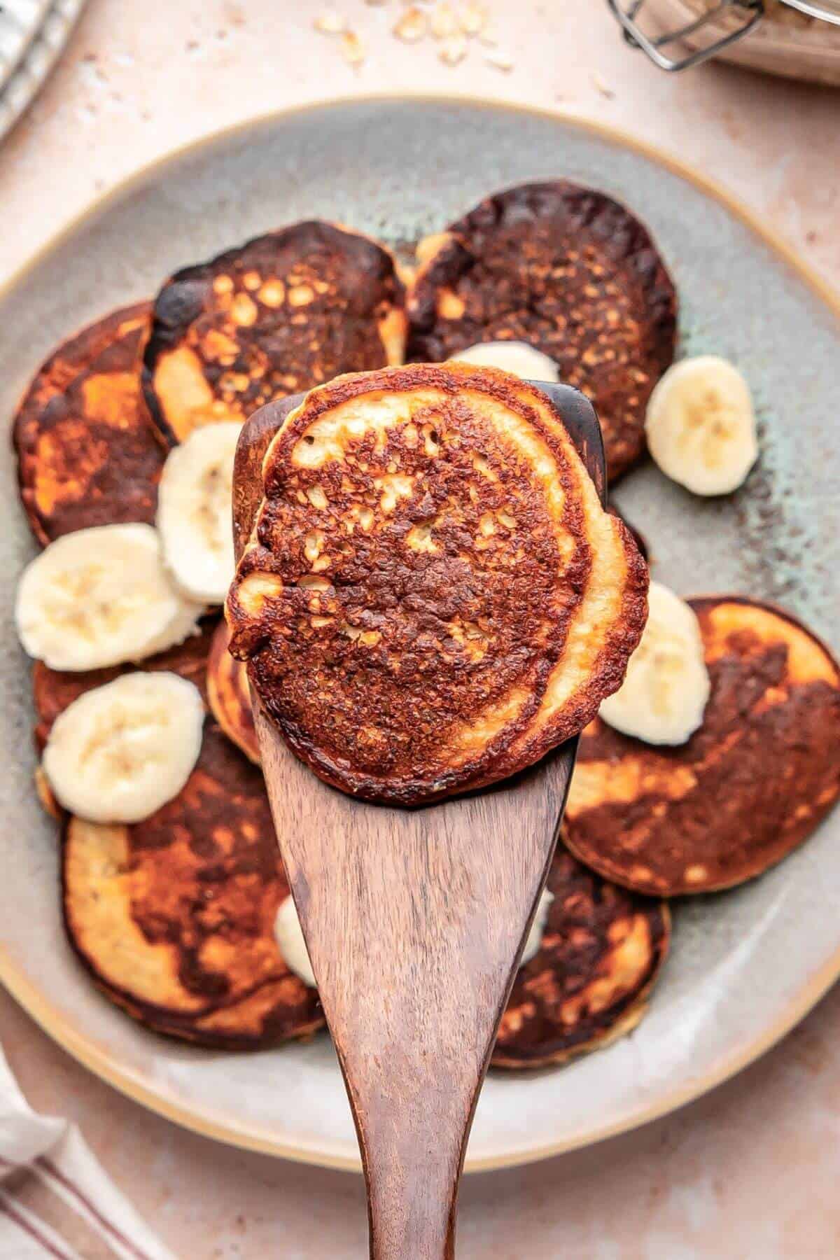 A wooden spatula holds up a browned pancake above a plate of pancakes garnished with banana slices.