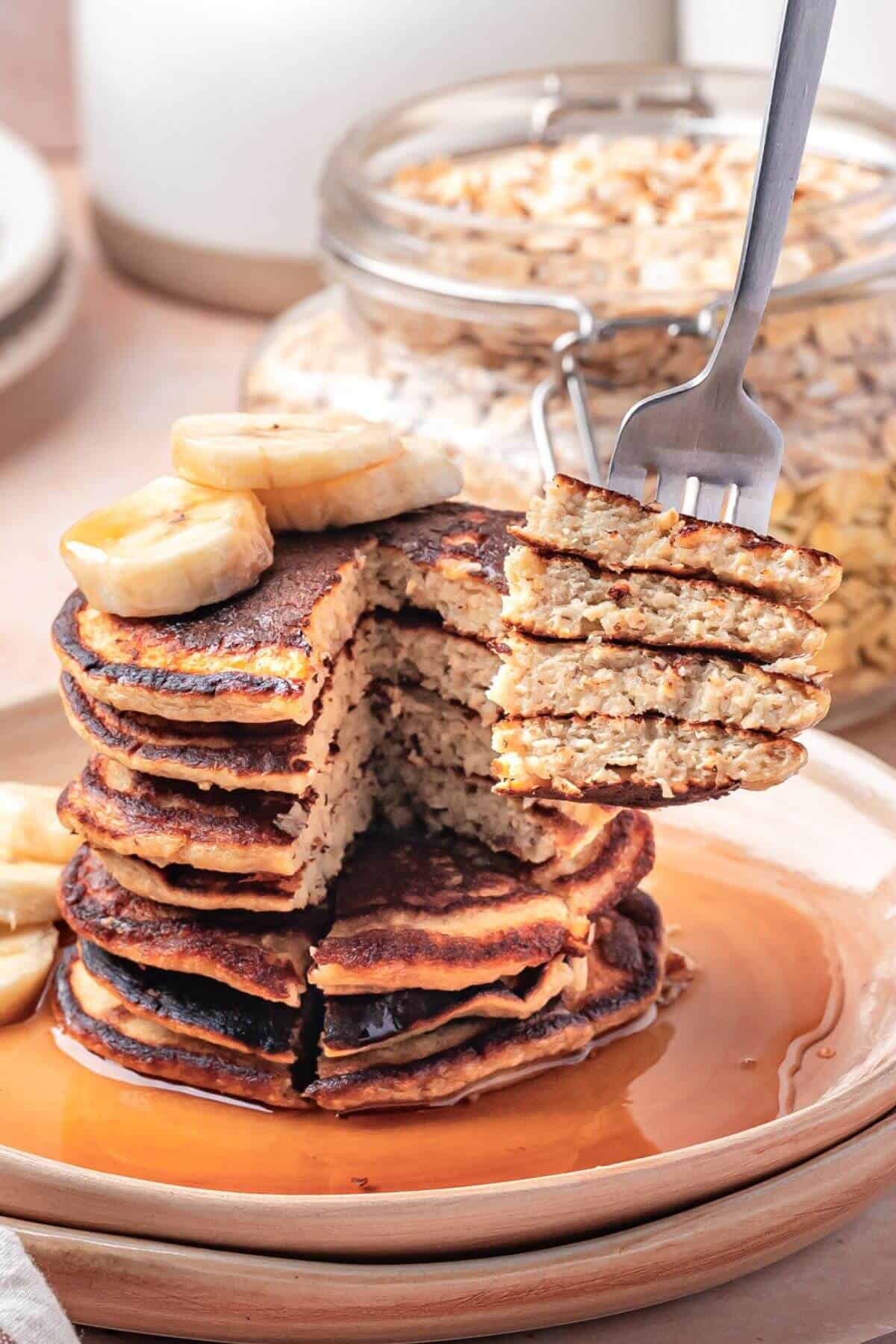 A stack of pancakes topped with banana slices sits on a plate with syrup, while a fork holds a cut section of pancakes above the stack.