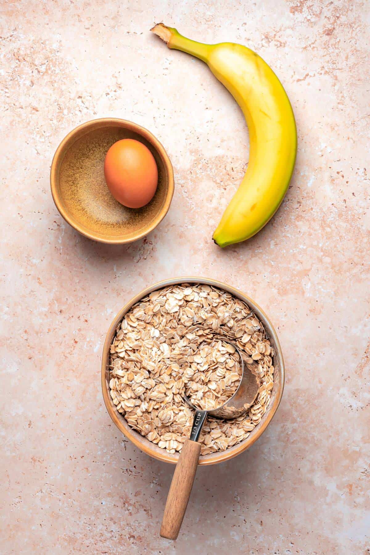 A banana, a bowl with a brown egg and brown sugar, and a larger bowl of rolled oats with a wooden scoop are arranged on a light countertop.