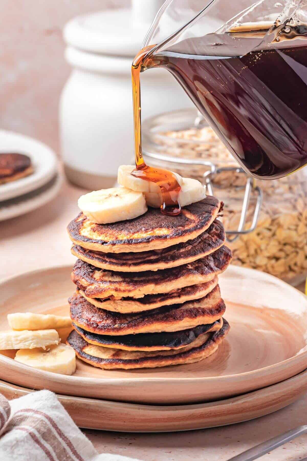 A stack of pancakes topped with banana slices is being drizzled with syrup from a glass pitcher, with extra banana pieces on the plate.