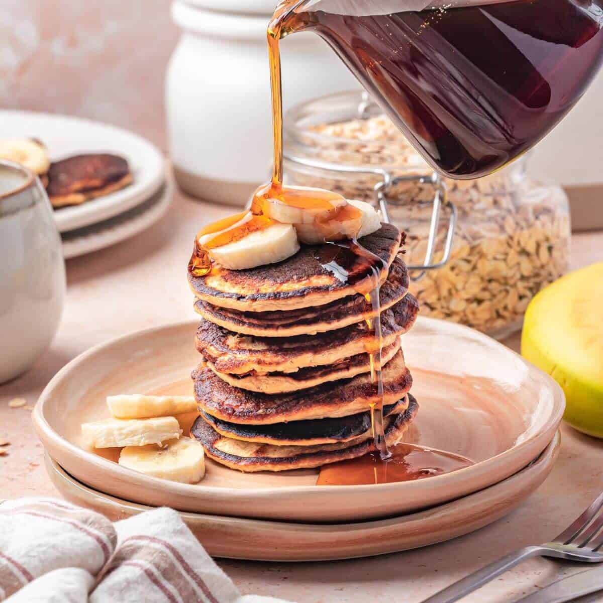 A stack of pancakes topped with sliced bananas is being drizzled with syrup, with more banana slices on the side and jars of oats in the background.