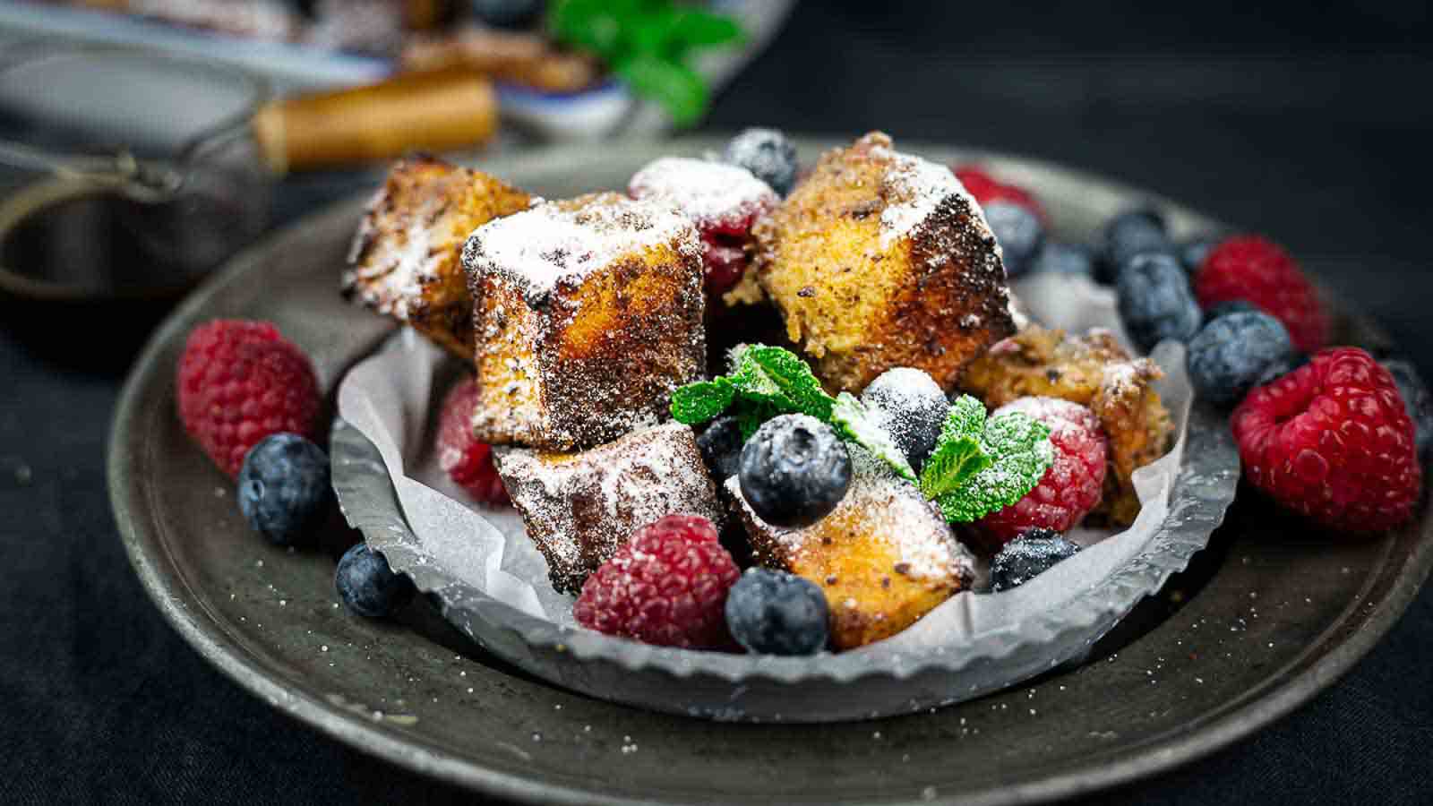A plate with cubes of French toast dusted with powdered sugar, garnished with fresh raspberries, blueberries, and a sprig of mint.