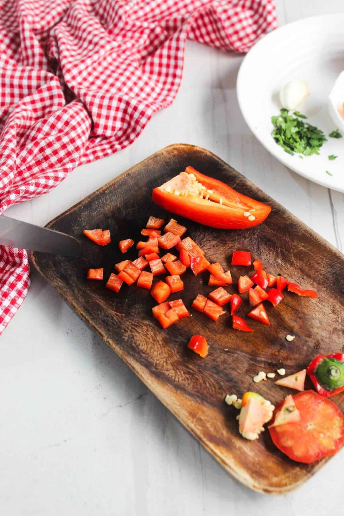 Diced red bell pepper on a wooden cutting board with a knife, next to a red checkered cloth and a plate with herbs and garlic.