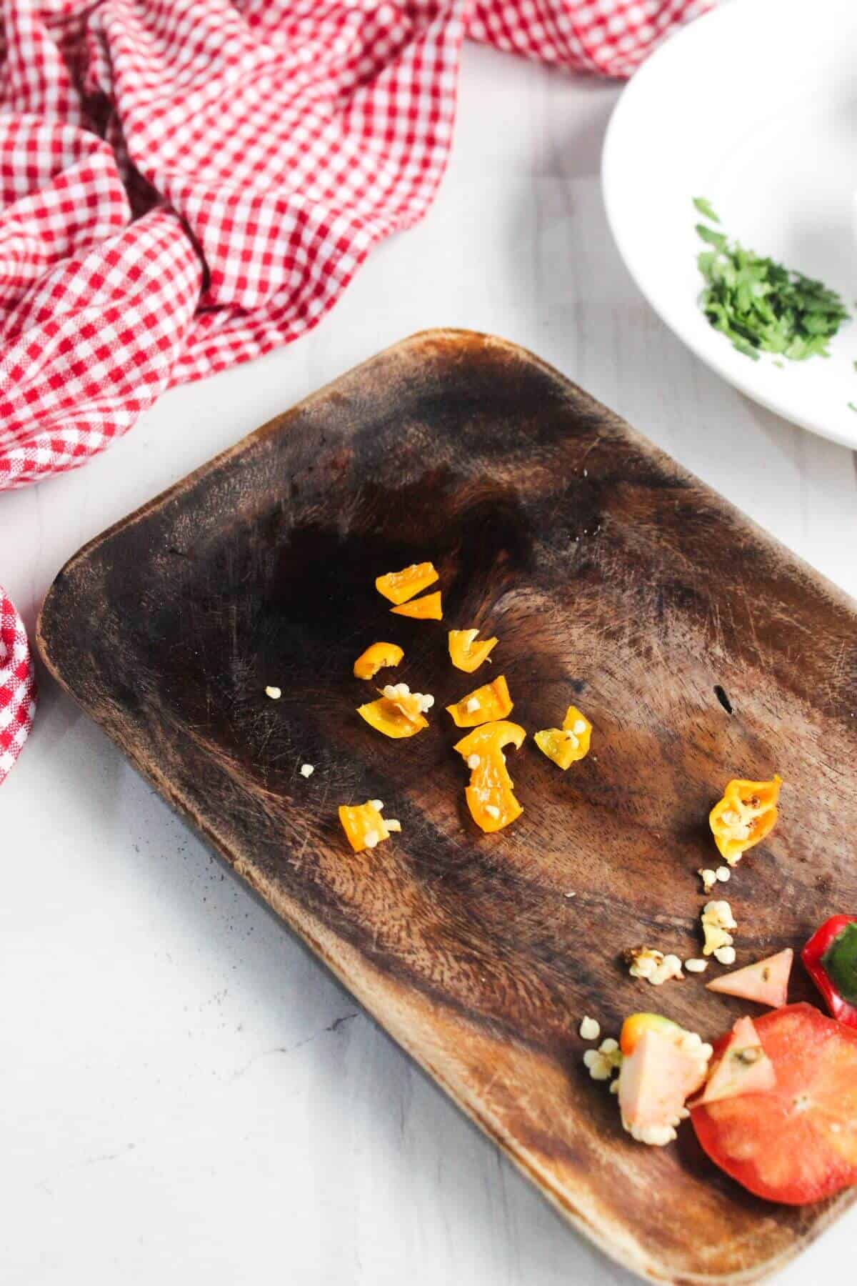 A wooden tray with chopped yellow chili pieces and seeds, beside a red checkered cloth and a white plate with herbs.
