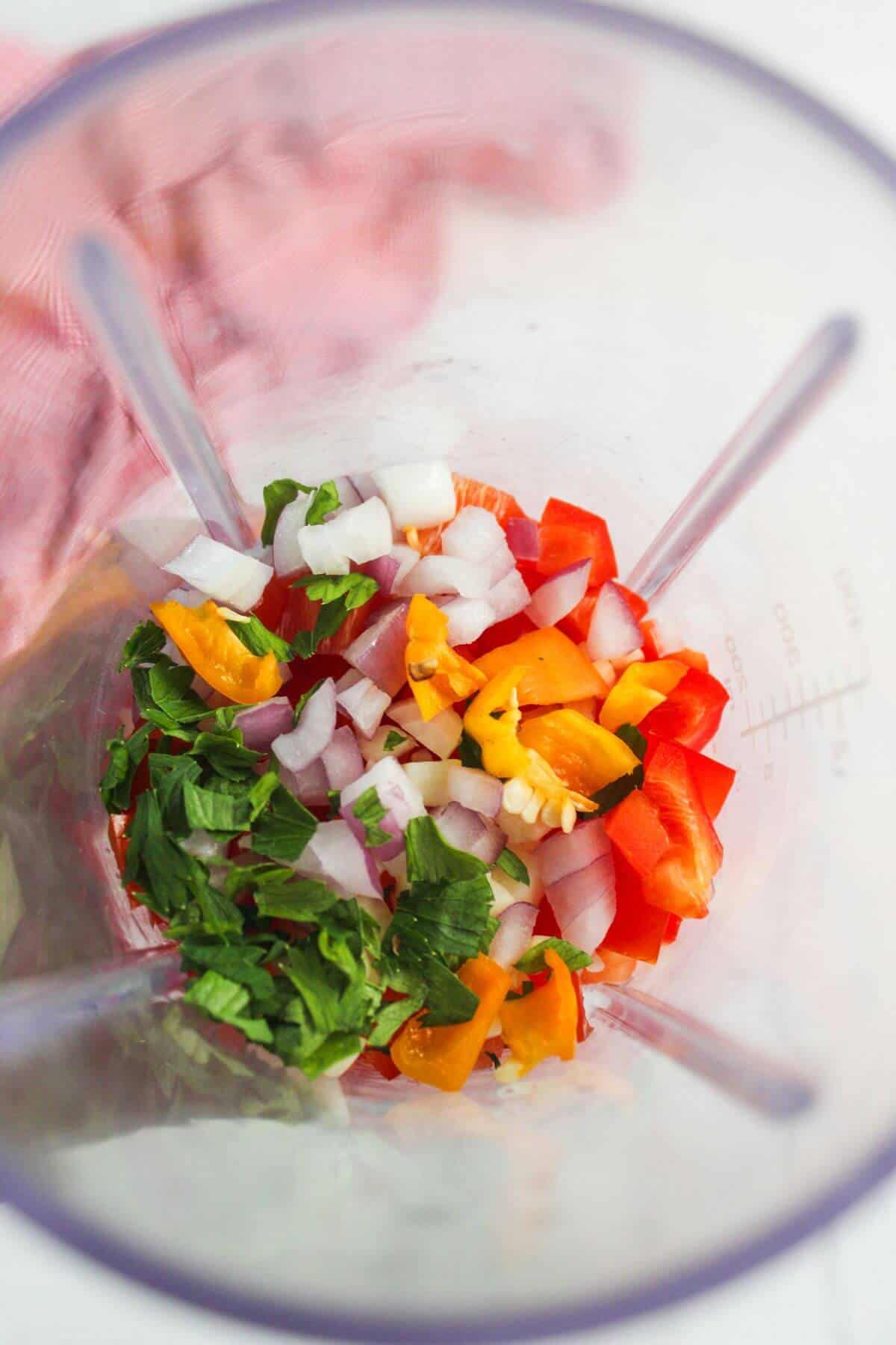 Chopped onions, bell peppers, and herbs in a blender jug, ready to be blended.