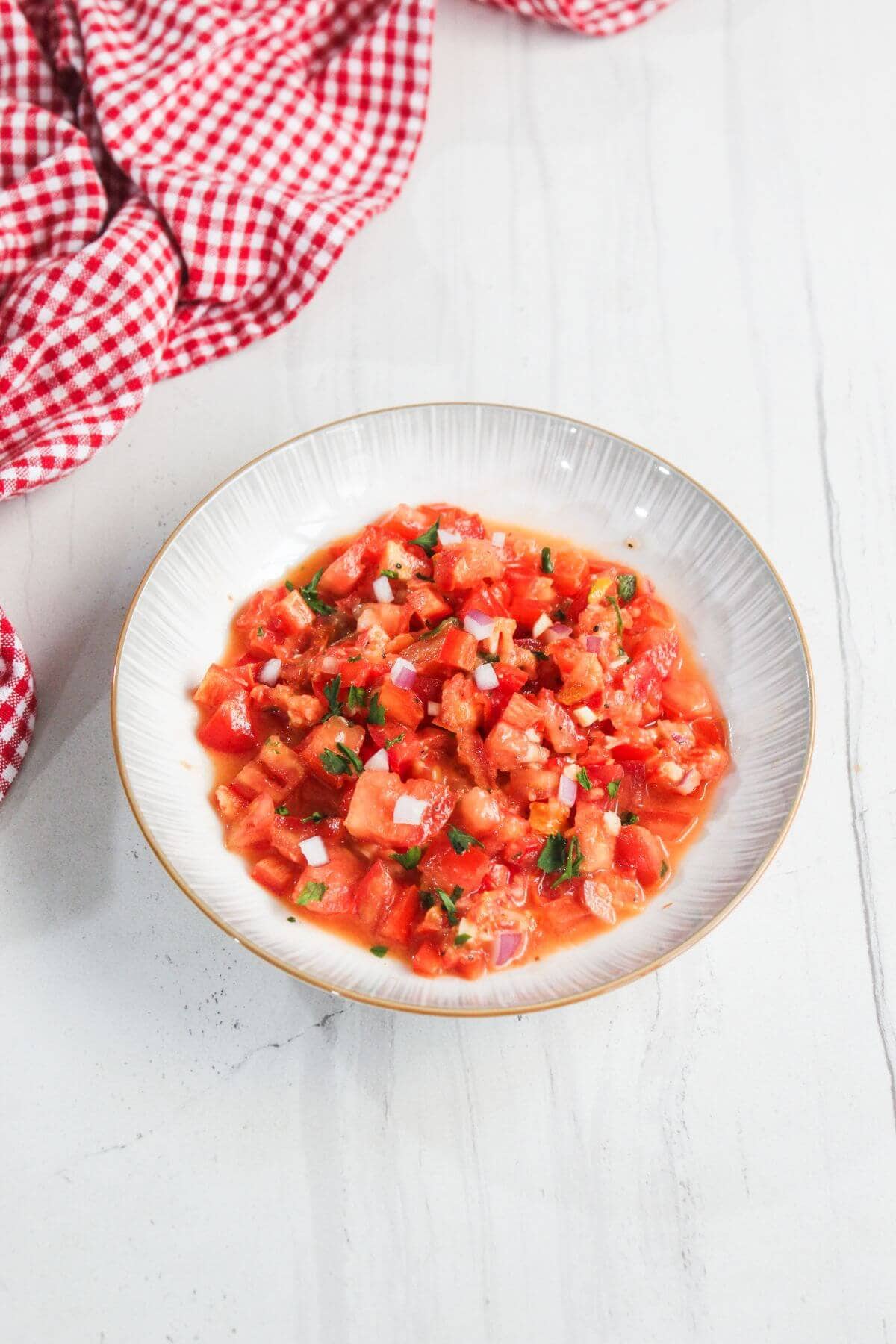 A white bowl filled with chopped tomatoes and herbs sits on a white surface next to a red and white checkered cloth.