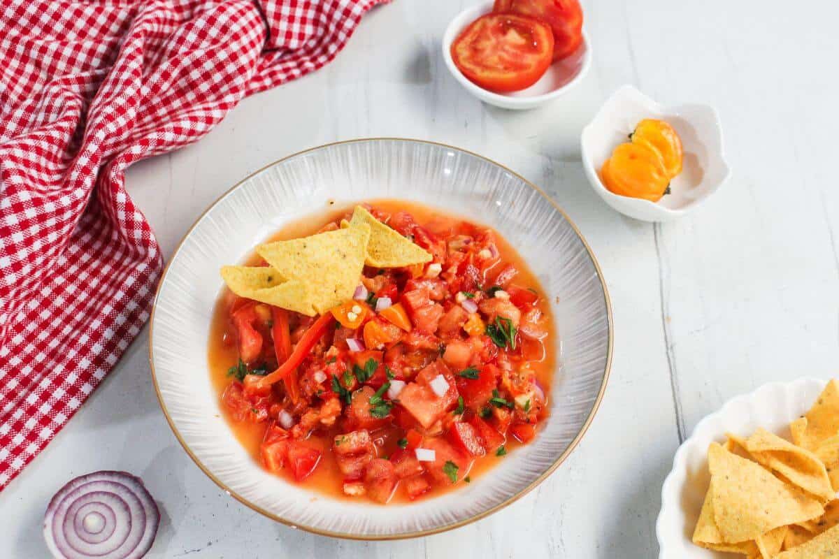 A bowl of tomato salsa with chopped onions and herbs, garnished with tortilla chips, sits on a white surface next to sliced tomatoes, peppers, and a red checkered cloth.