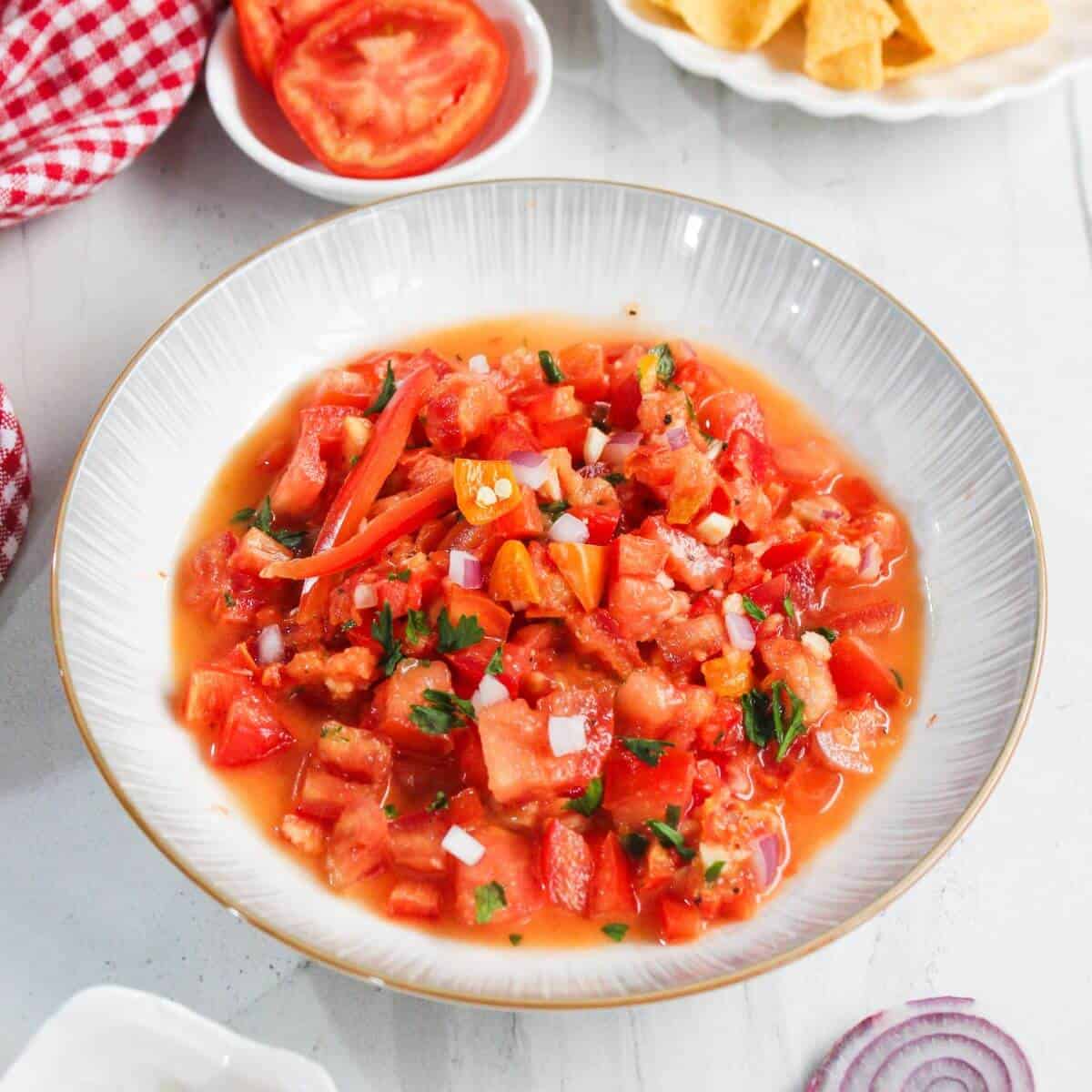 A bowl of fresh salsa with chopped tomatoes, onions, cilantro, and peppers sits on a white table, surrounded by tortilla chips and sliced tomatoes.