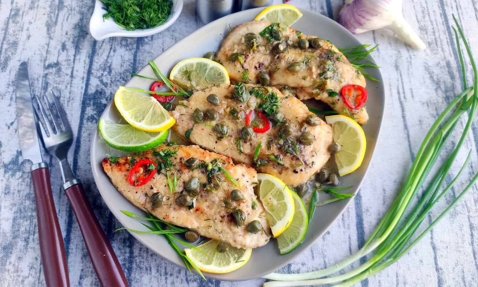 Plate of cooked chicken breasts garnished with capers, lemon slices, red chili, and fresh herbs, served with green onions on a rustic wooden table.
