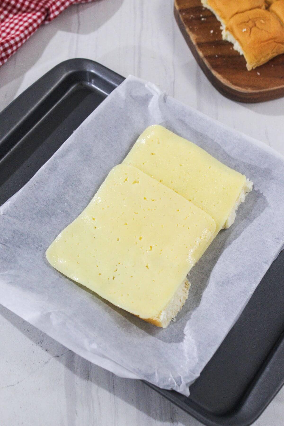 Two rectangular pieces of bread with a layer of yellow cheese on top are placed on a parchment-lined baking tray.