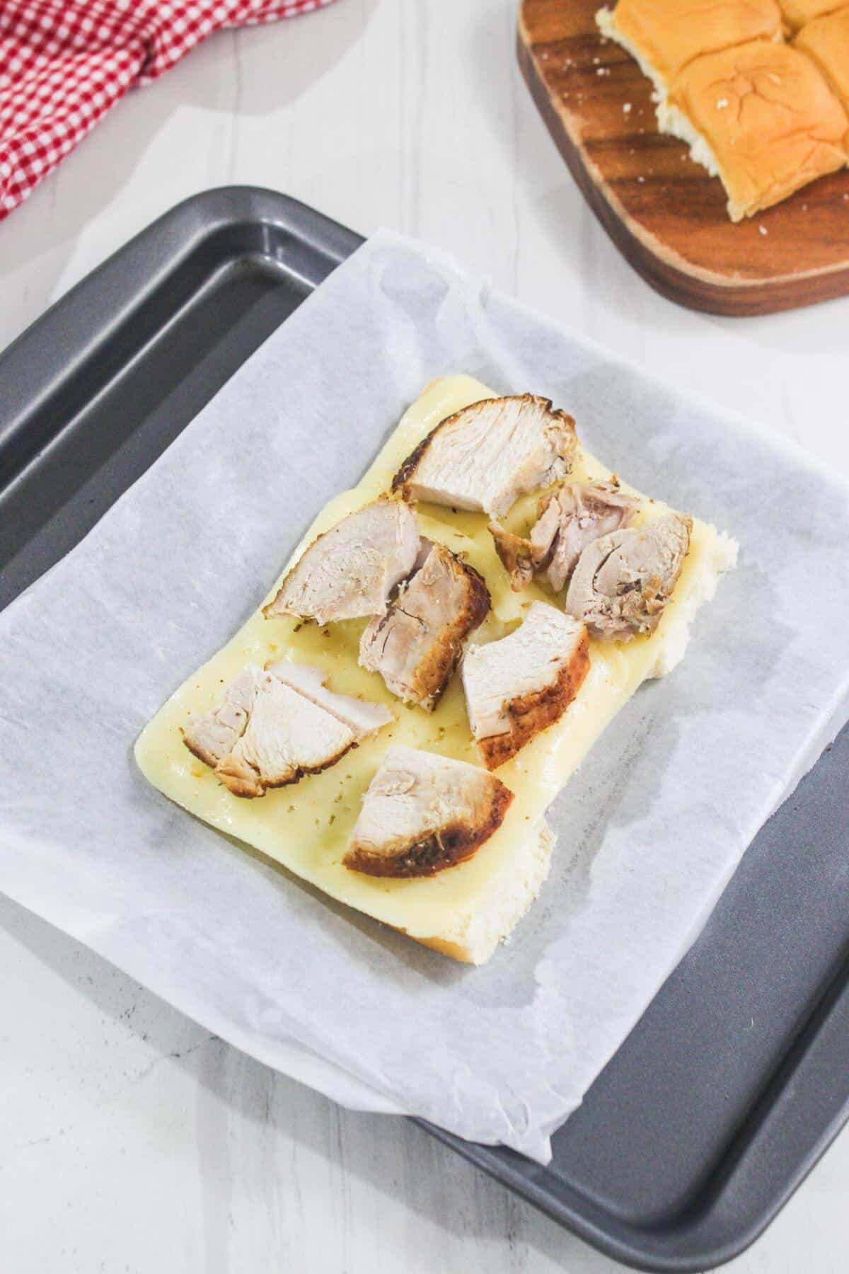 A slice of bread topped with melted cheese and pieces of cooked chicken rests on parchment paper in a baking tray.