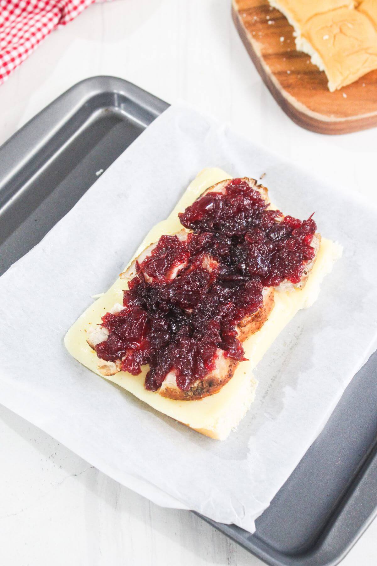A sandwich roll on parchment paper is topped with sliced sausage, melted cheese, and a layer of dark red sauce or relish.