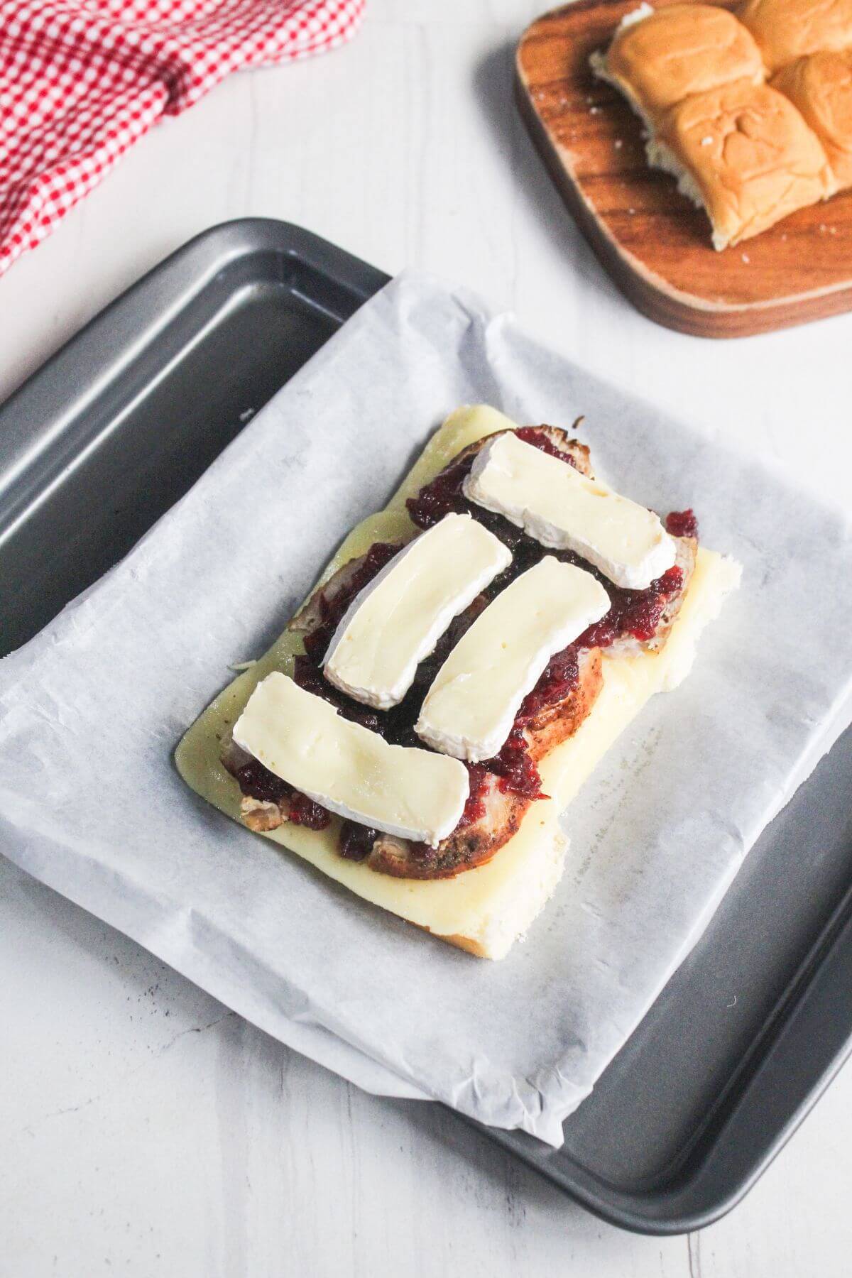 An open sandwich roll topped with cranberry sauce and slices of brie cheese on a parchment-lined baking tray, with extra rolls in the background.