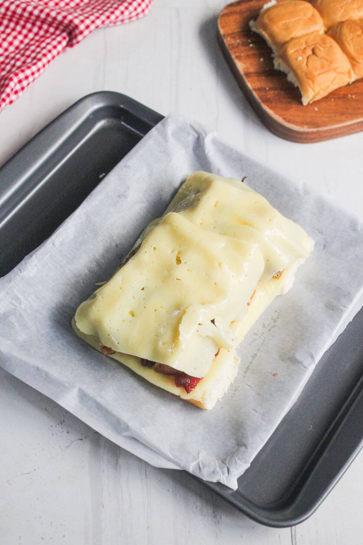 A sandwich covered with melted cheese sits on parchment paper in a baking tray, with a partially visible tray of slider buns in the background.