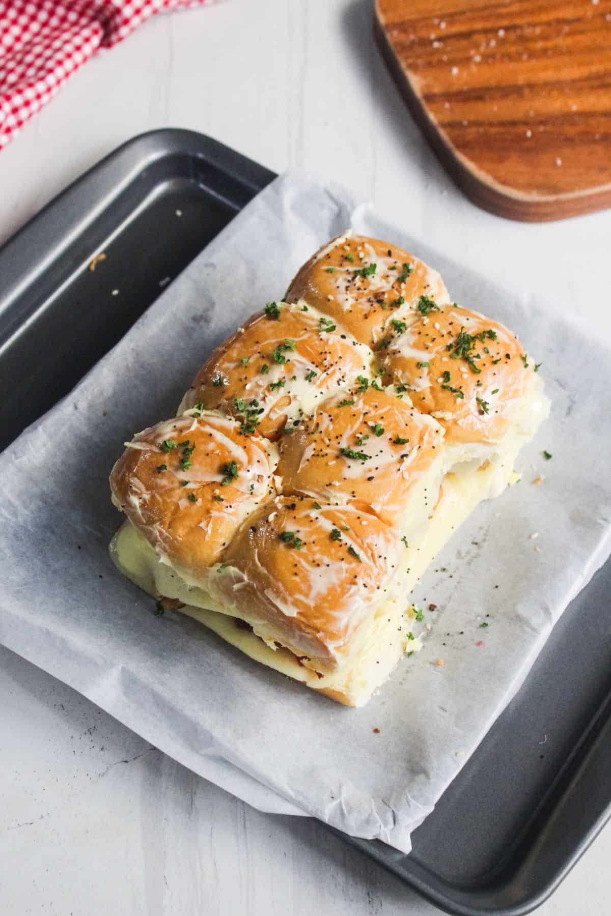 Six baked slider sandwiches with melted cheese and herbs on top, arranged on parchment paper in a baking tray.