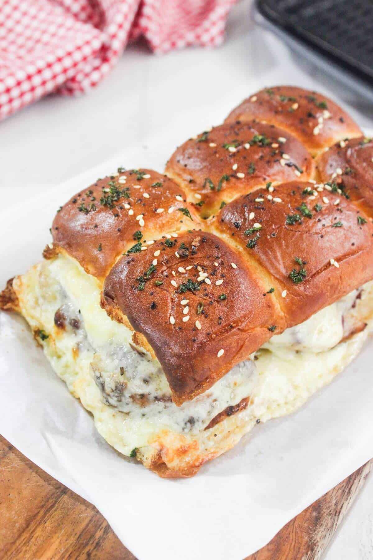 A close-up of cheesy meatball sliders on a wooden board, featuring golden brown buns topped with herbs and sesame seeds.