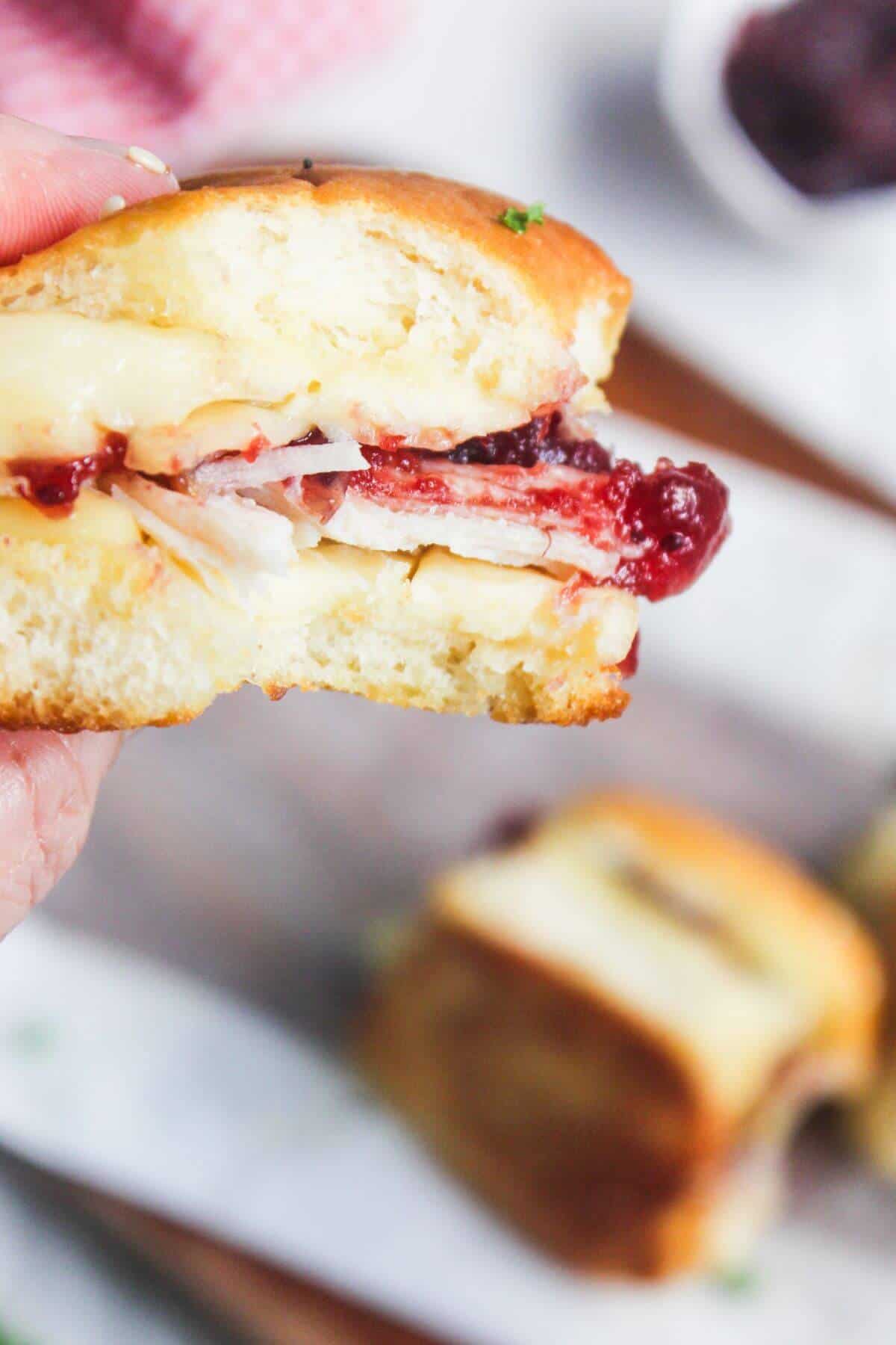 A hand holding a sandwich with turkey, cheese, and cranberry sauce on a soft roll, with another sandwich blurred in the background.