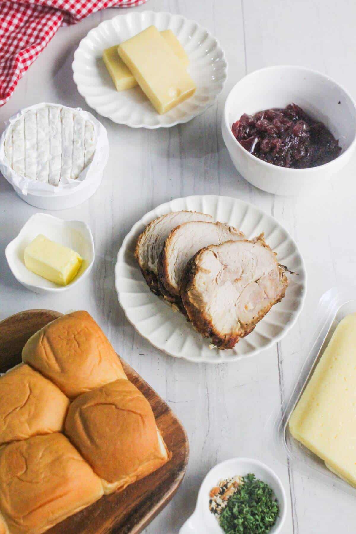 A table with sliders, sliced roast pork, cheese, butter, cranberry sauce, herbs, and spices arranged in separate dishes on a white surface.