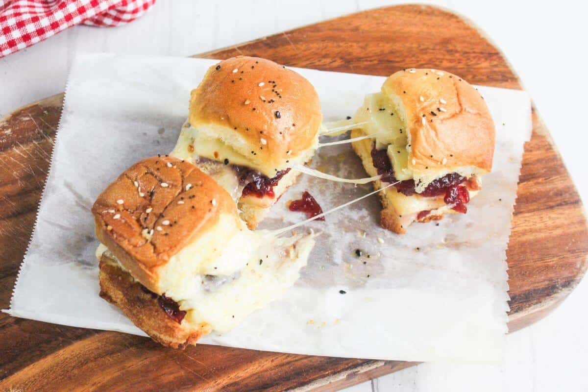Three slider sandwiches with melted cheese and a red filling on parchment paper atop a wooden cutting board.