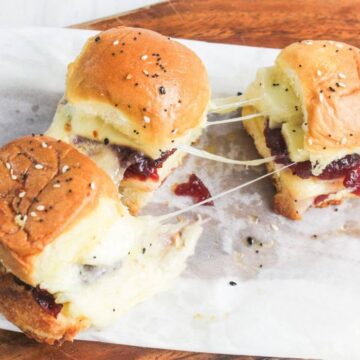 Three cheesy sliders with sesame seed buns, melted cheese, and sauce are placed on parchment paper atop a wooden board.