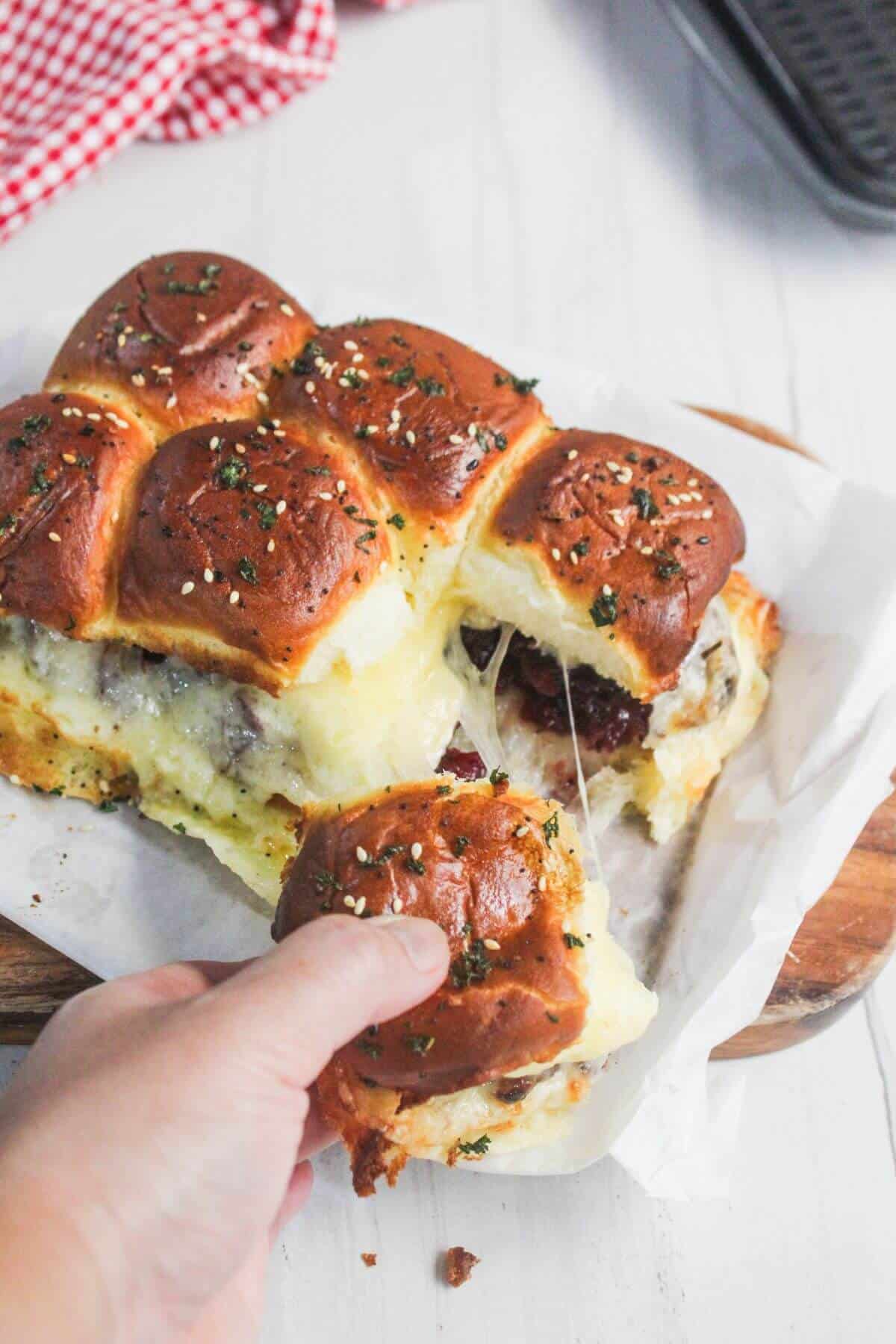 A hand pulls apart a cheesy slider from a batch of baked sliders on parchment paper, with melted cheese stretching between the buns.