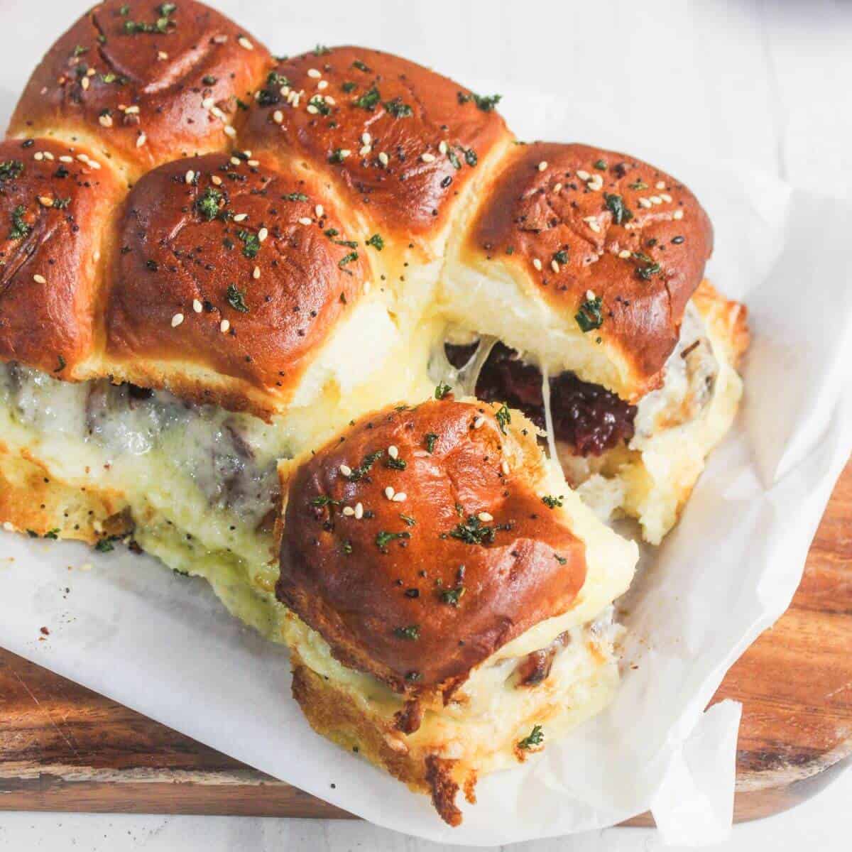 A tray of golden-brown baked sliders with melted cheese and herbs on top, one slider partially pulled away, on parchment paper and a wooden board.