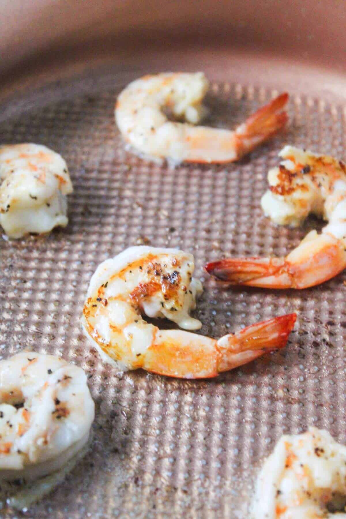 Six shrimp are being cooked in a pan, with some lightly browned and others still partially raw.