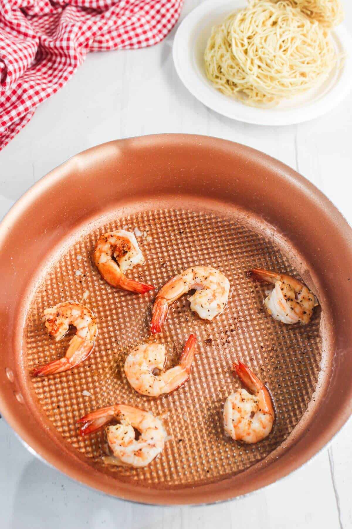 Cooked shrimp in a copper skillet with seasoning, with a plate of uncooked noodles and a red checkered cloth in the background.