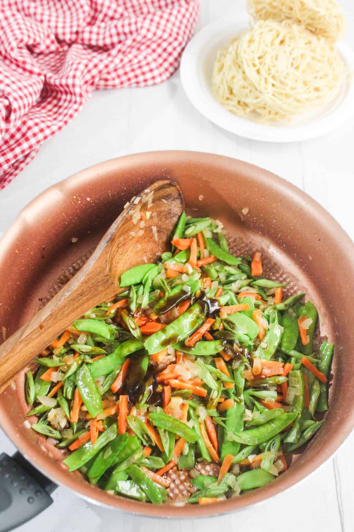 Chopped snow peas, carrots, and onions being sautéed in a copper pan with a wooden spoon; a plate of uncooked noodles is in the background.