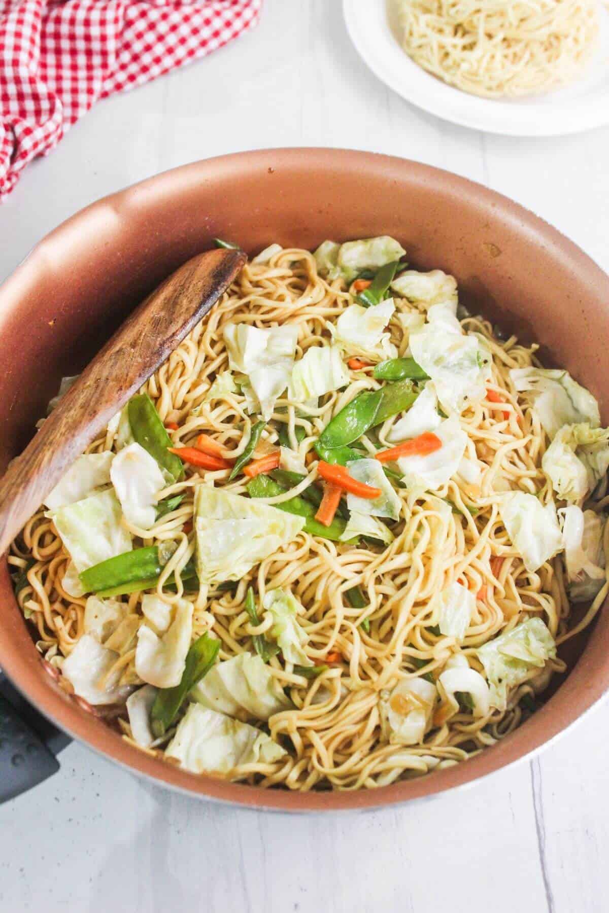 A pan of cooked noodles mixed with chopped cabbage, snow peas, and carrots, with a wooden spatula resting on the side.