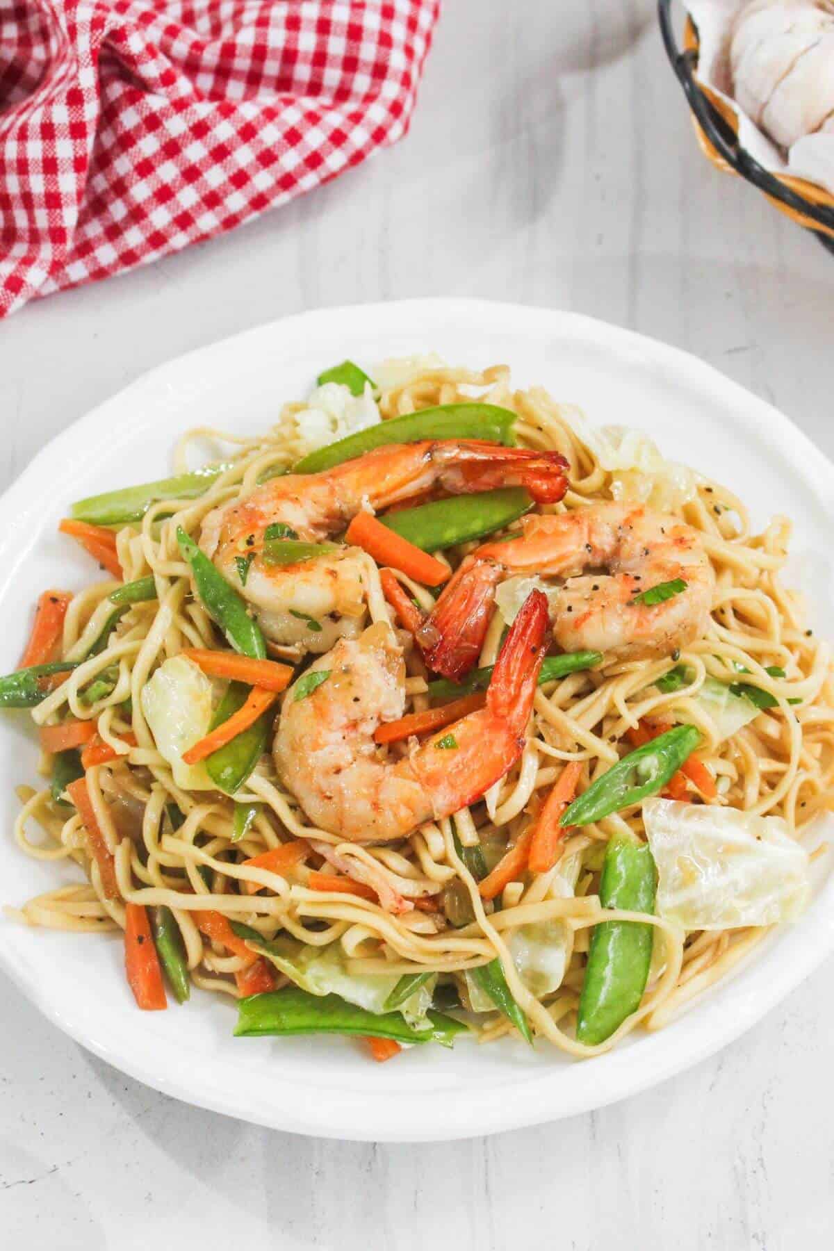A white plate with shrimp lo mein, noodles, shrimp, carrots, snow peas, and cabbage on a white surface with a red checkered cloth.