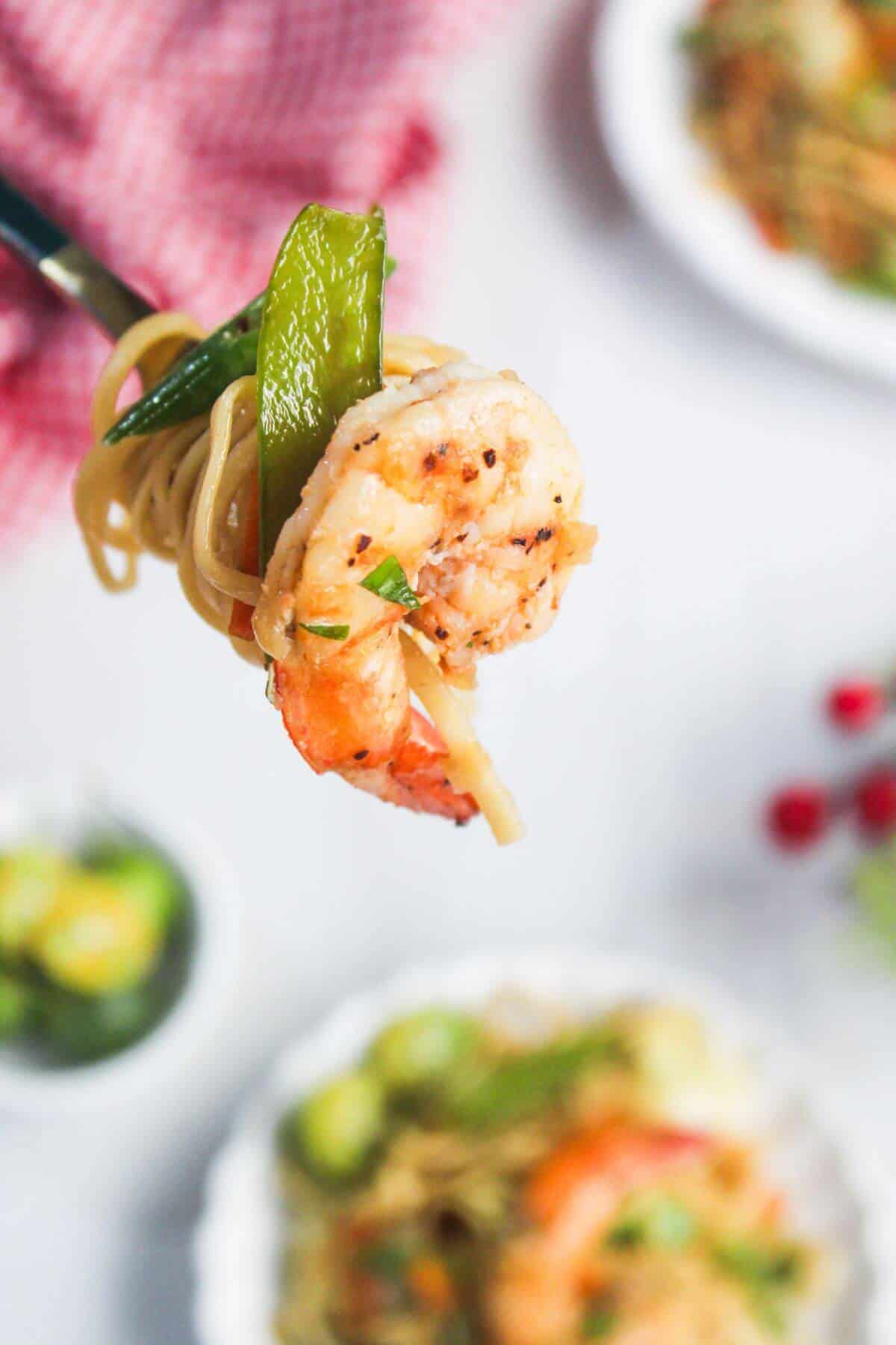 A fork holds a bite of noodles, shrimp, and a green vegetable above a plate of food on a white surface.