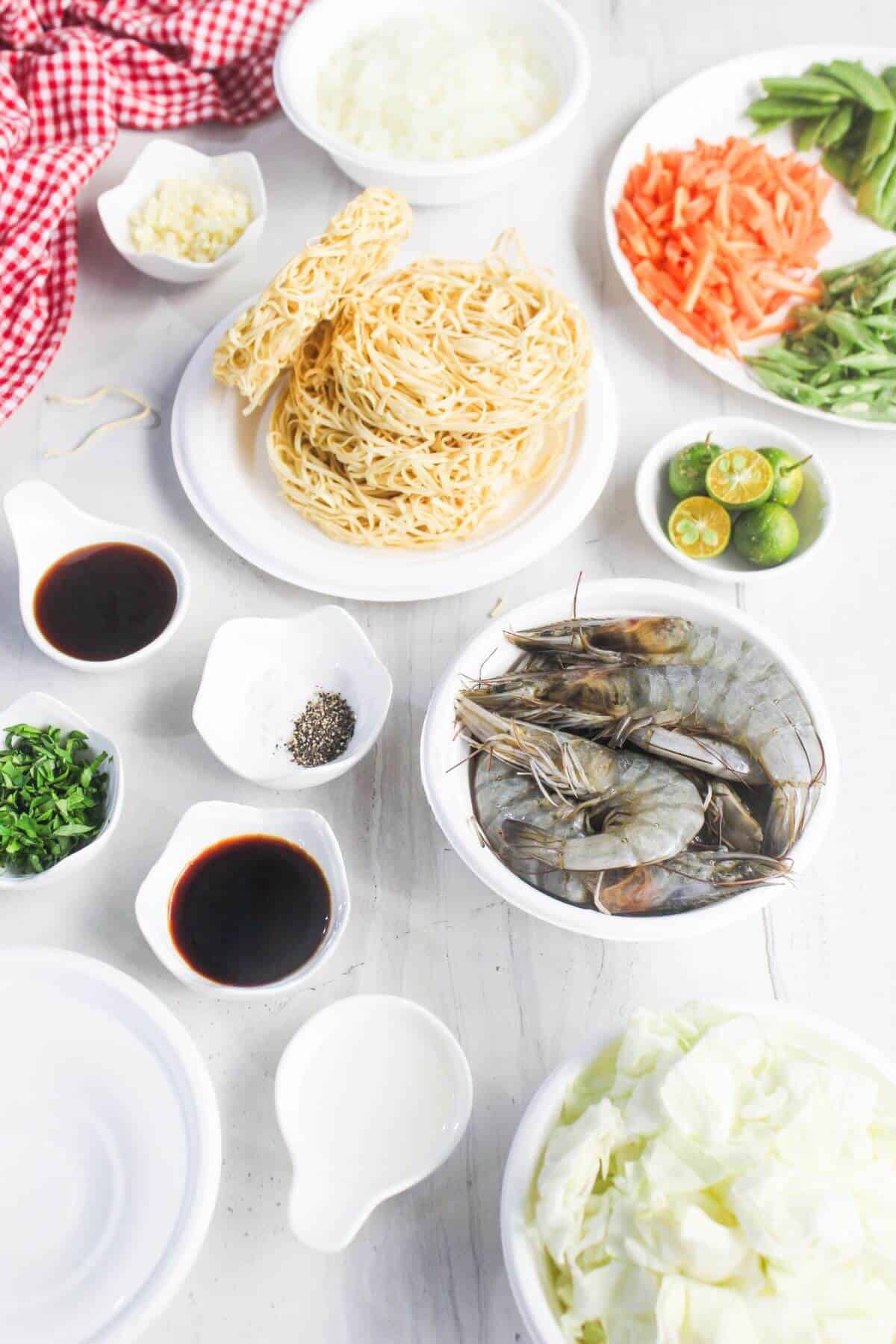 Various ingredients for a noodle dish are arranged on a white surface, including raw shrimp, noodles, vegetables, seasonings, and sauces in bowls and plates.